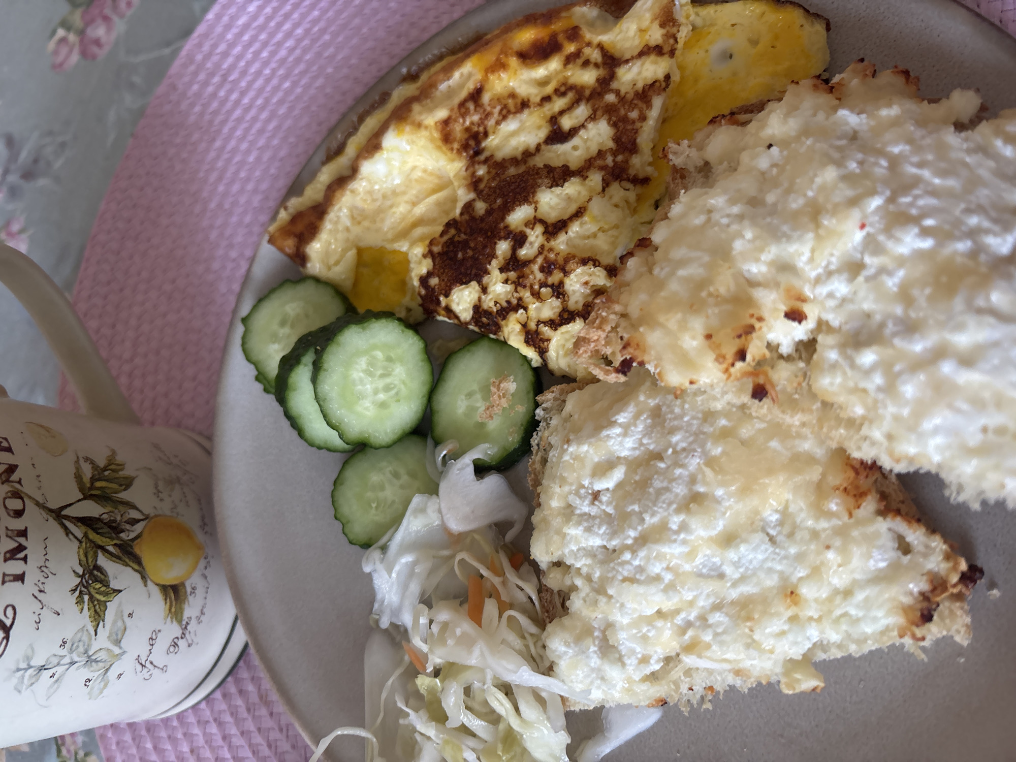 A close-up shot shows a breakfast plate with food and a mug in the background. The plate contains two pieces of bread topped with a white, fluffy substance, possibly cottage cheese or a similar spread, with some browned edges indicating it may have been toasted or baked. To the right of the bread is a folded omelet with browned areas. In the upper left quadrant of the plate is a small portion of shredded cabbage and carrot salad. Several slices of cucumber are arranged next to the salad and omelet. In the background, a mug with a decorative design, possibly featuring a lemon branch, rests on a pink placemat. The overall setting appears to be a home or cafe, with a blurred background that suggests an indoor environment. There are no individuals present in the image. The scene is consistent with a breakfast meal. The location of this scene in Naberezhnye Chelny, Russia, cannot be determined from the visual information provided.