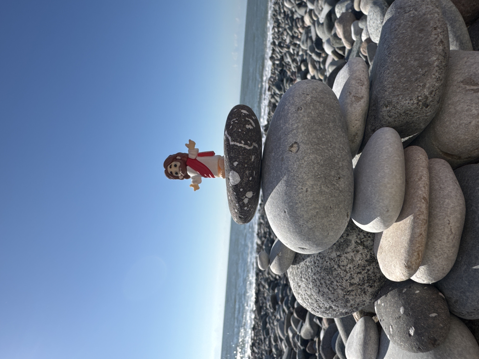 A small figurine of Jesus, wearing a white robe with a red sash, stands atop a stack of smooth, grey, and speckled rocks on a pebble beach. The ocean is visible in the background, with gentle waves meeting the shore under a clear blue sky. The scene is likely the Batumi Boulevard in Batumi, Georgia, a popular seaside promenade.
