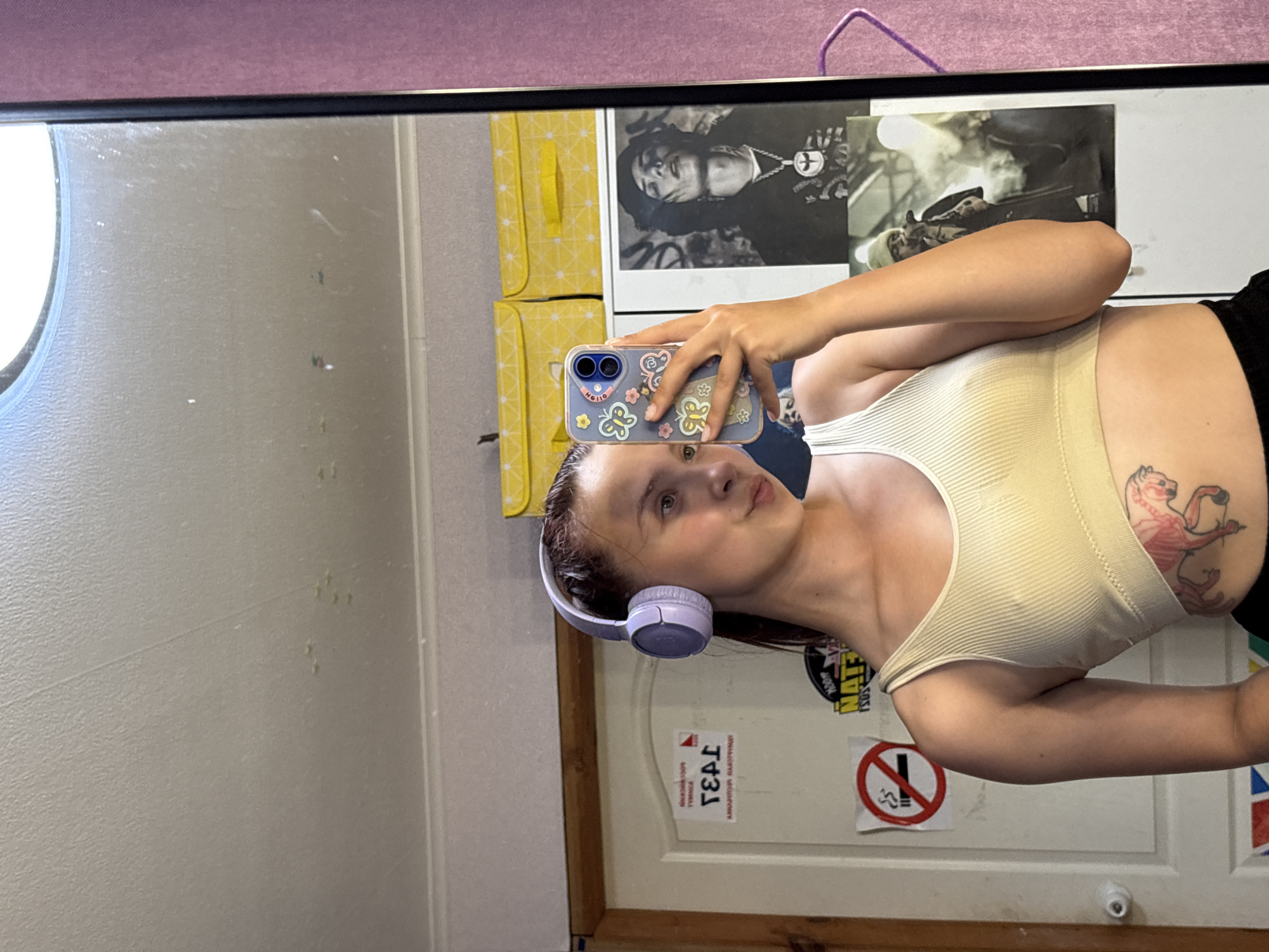 A woman wearing headphones and a tank top is taking a selfie in front of a mirror. Behind her, there is a poster, yellow storage boxes, and stickers on a white door, including a no-smoking sign. The location is not discernible from the image.