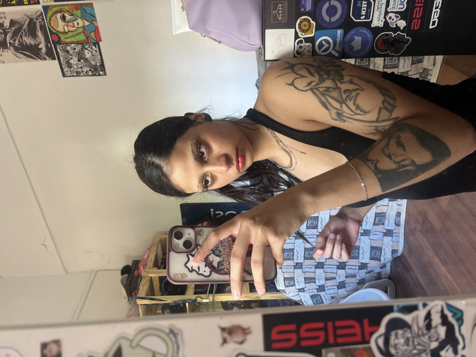 A young woman with dark hair and eyebrow piercings takes a mirror selfie. She holds her phone with a patterned case, her arms bearing tattoos. She is wearing a black top. Behind her, a room contains posters on the wall, a lavender bag, and a sticker-covered object. A checkered surface and fan are visible below. The camera captures her from a tilted angle.