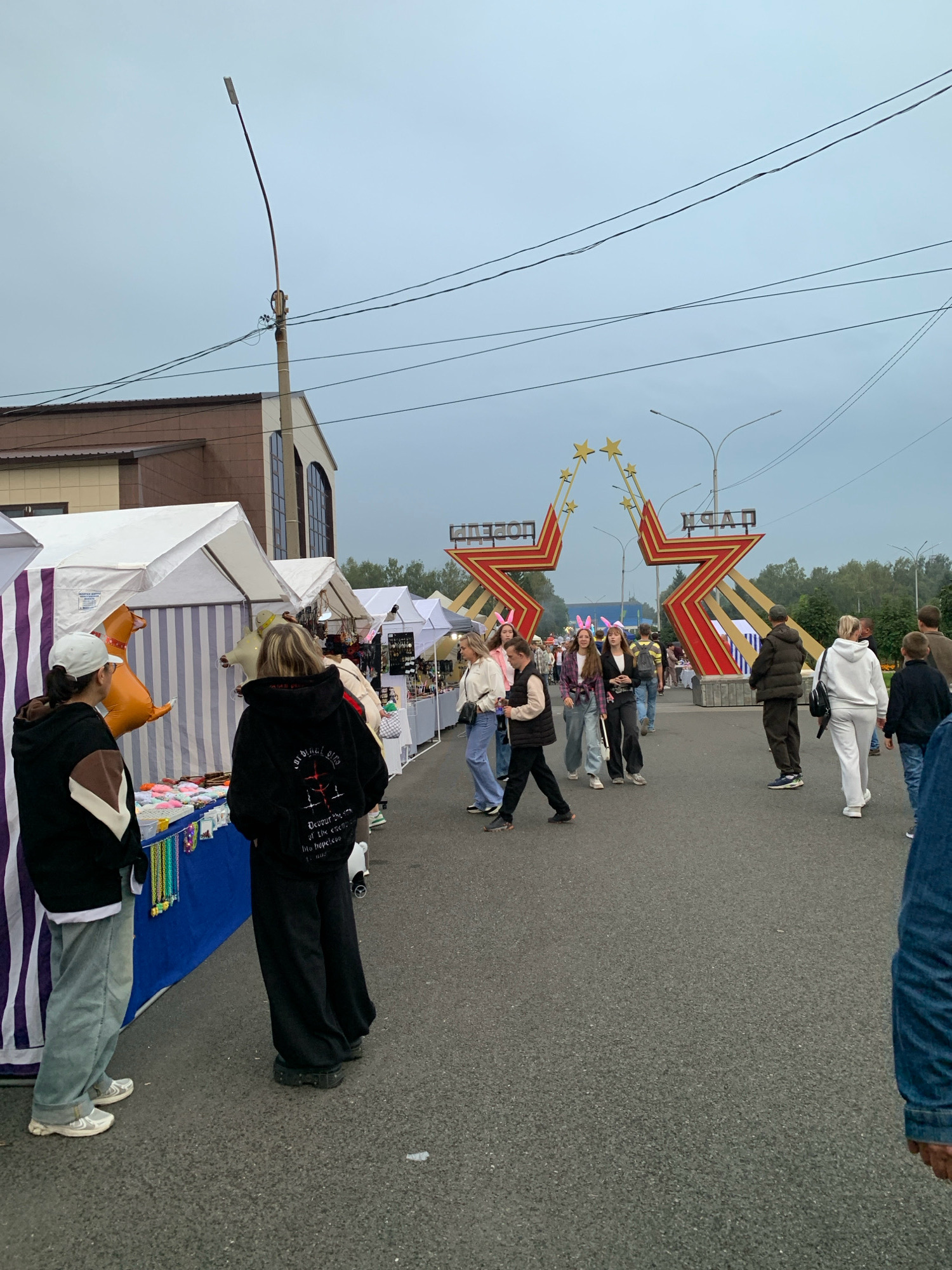 A street fair is taking place under an overcast sky in Prokopyevsk, Russia. A large, red and gold star-shaped archway with the words "ПОБЕДА" (Pobeda, meaning "Victory") and "ПАРК" (Park) is visible in the background. Numerous people are walking along an asphalt path, with market stalls set up on either side. Some stalls have white tents with colorful striped fabric. On display at one stall are various colorful beaded necklaces and bracelets. People of various ages are present, some are wearing casual clothing, while others are dressed in hoodies and jeans. A few individuals are sporting bunny ear headbands. The overall atmosphere suggests a community gathering or event.