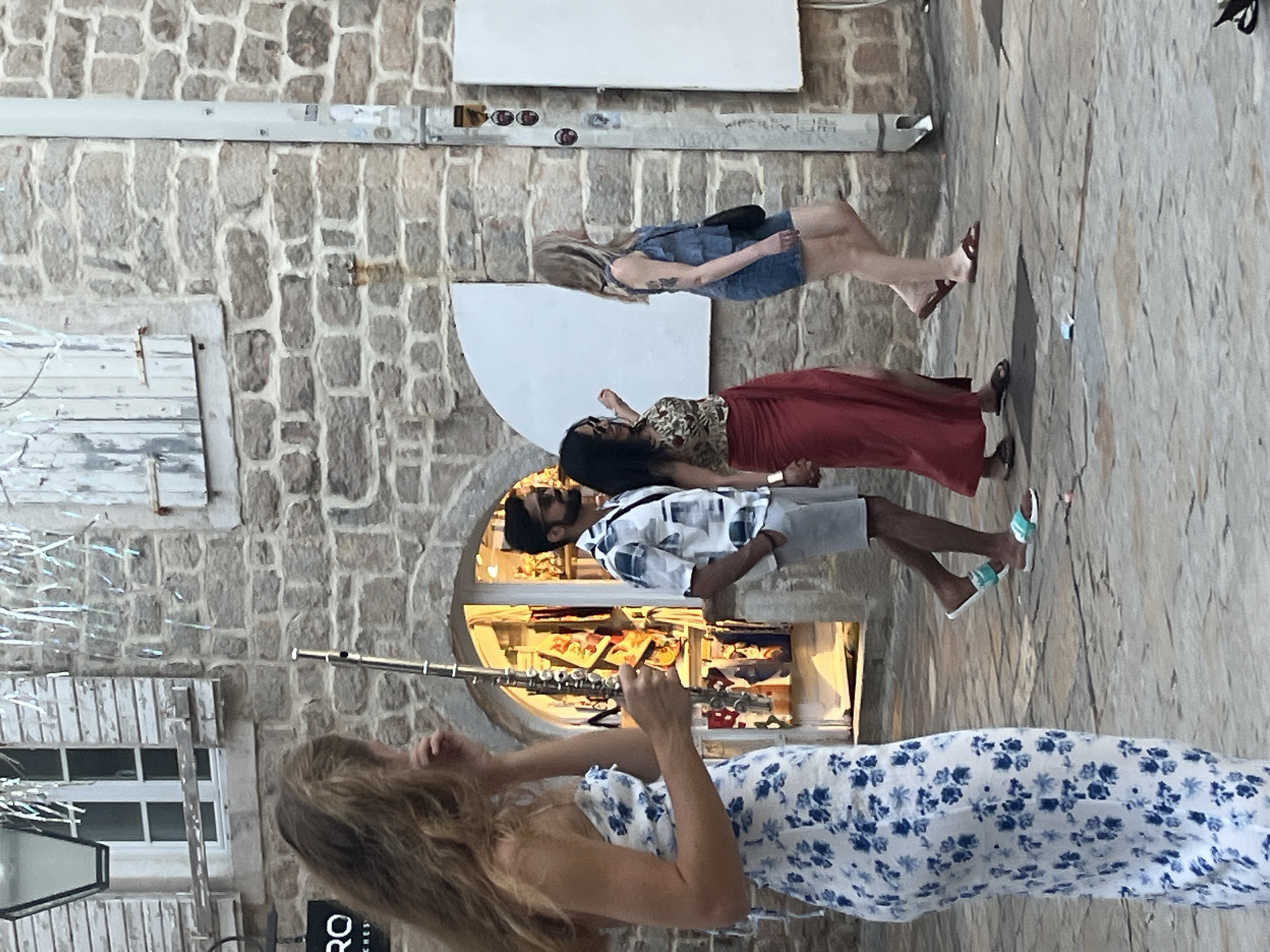 The image shows a street scene in Budva, Montenegro. In the foreground, a woman in a blue and white floral dress is holding a flute. Behind her, a group of people is walking past a shop entrance. One man is wearing a patterned shirt and shorts, while a woman next to him wears a red skirt and a patterned top. Another woman in a denim dress walks behind them. The background features a stone building with wooden shutters and a light fixture. The ground is paved with cobblestones.