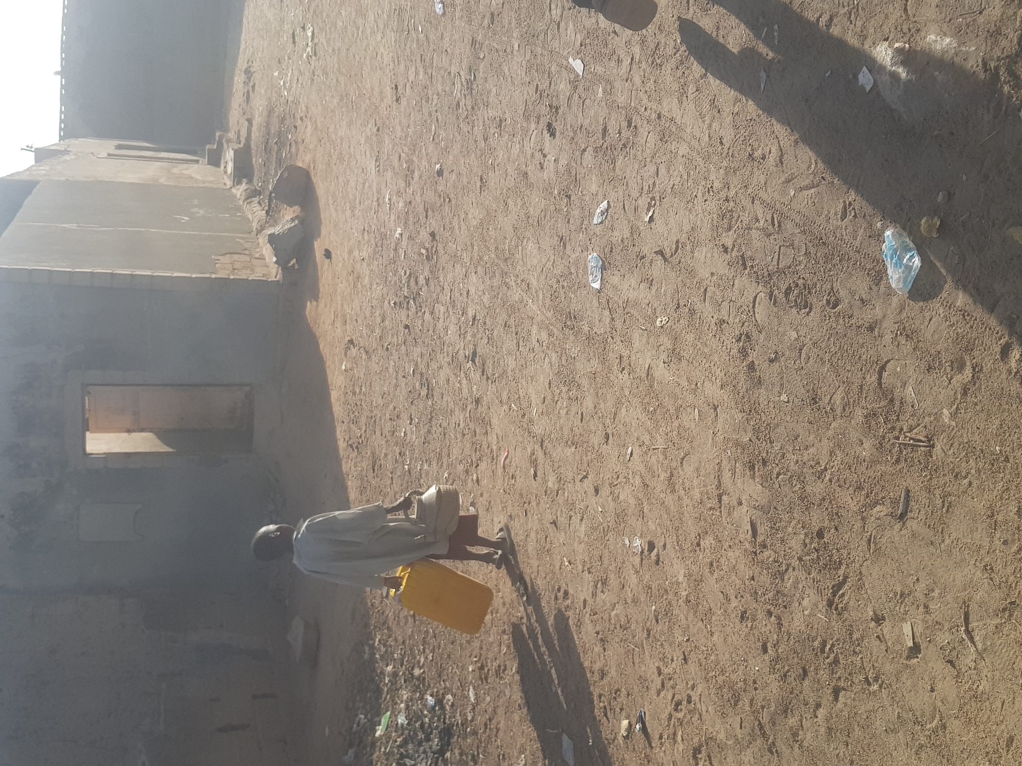A man walks along a dirt road in Dutsinma B, Nigeria. He carries a yellow jerrycan in his right hand and a tan-colored container on his head. The man is wearing a light-colored, long-sleeved shirt. The road is littered with scattered debris. An unfinished building with a visible window frame stands to the left of the road. The scene is illuminated by bright sunlight, casting shadows.