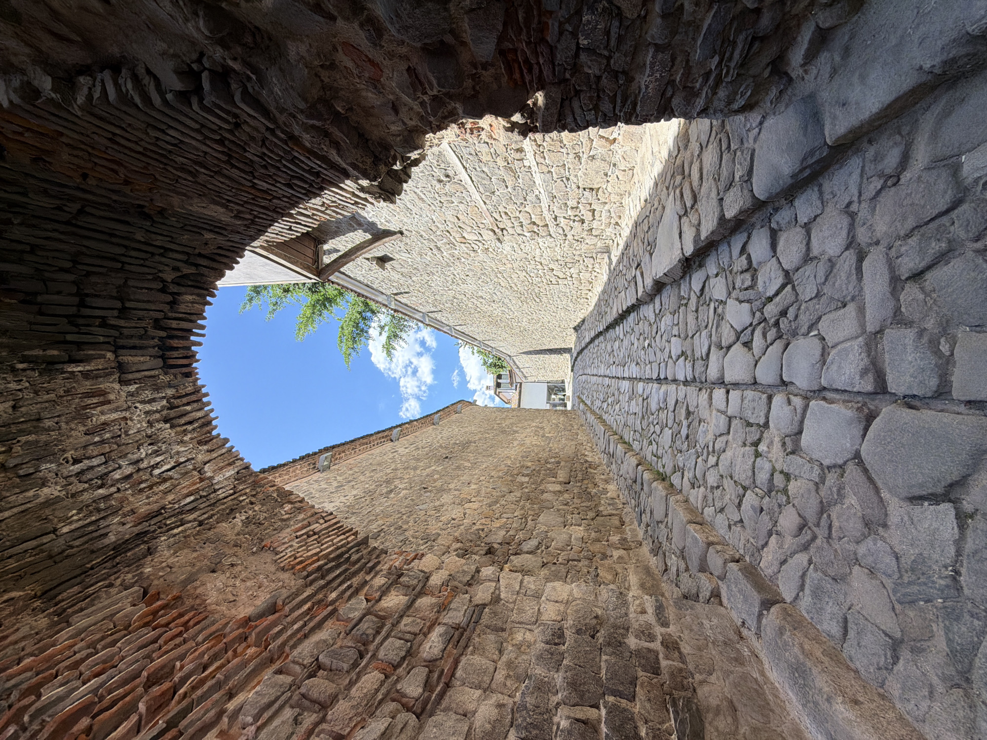 Low-angle shot of a narrow street in Plovdiv, Bulgaria, framed by old stone and brick walls. The street surface is paved with cobblestones, featuring a shallow drainage channel running down the center. Walls are constructed from mixed materials, including brick and roughly cut stone blocks. Above, a patch of blue sky is visible, punctuated by glimpses of green foliage and white clouds. The architectural details suggest a historical urban environment. No people are visible.