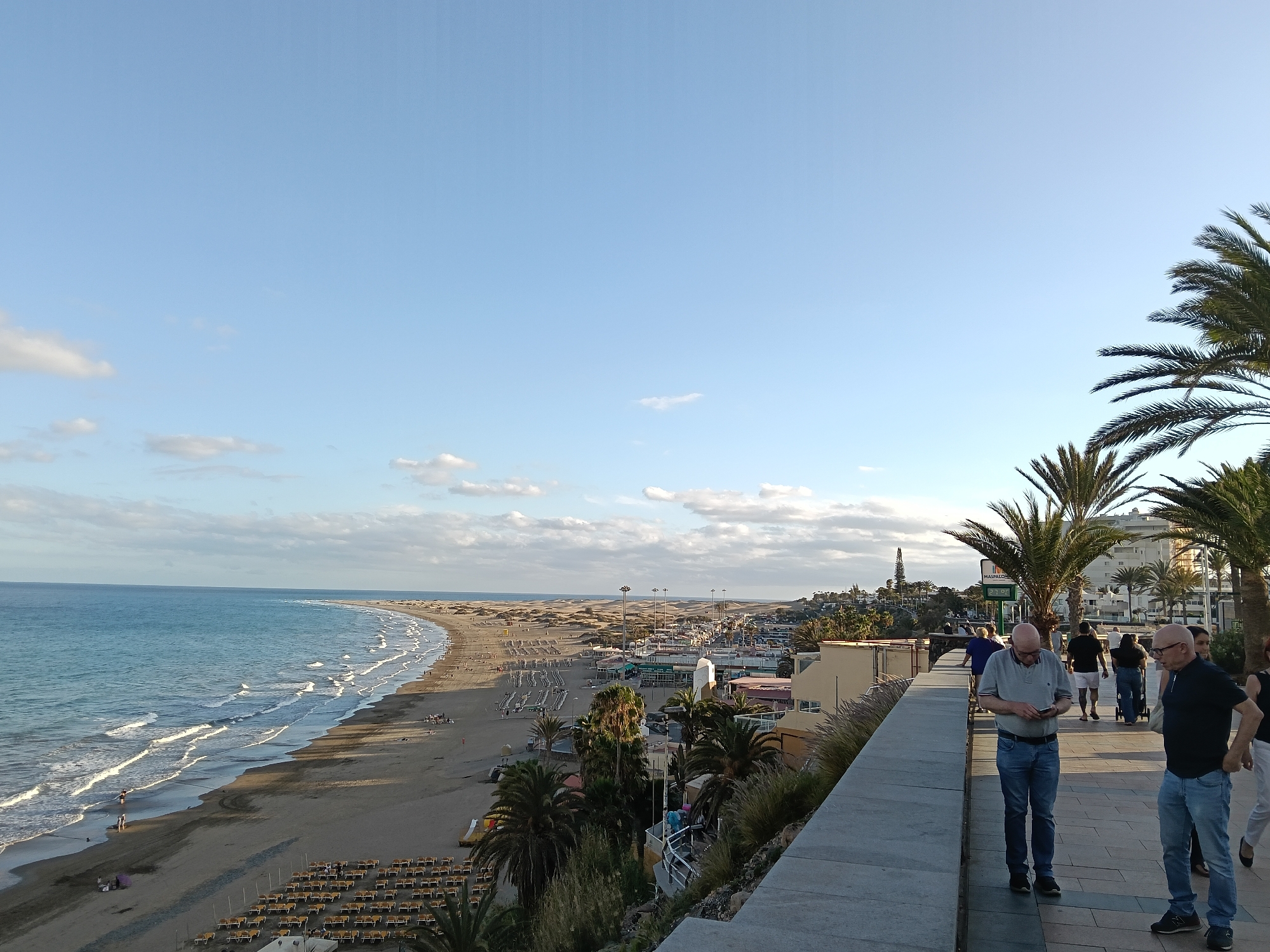 The image captures a sunny day at the beach in San Bartolomé de Tirajana, Spain. The scene overlooks a long sandy beach with sunbeds and sunbathers, bordered by the Atlantic Ocean with gentle waves. A row of buildings and vegetation separates the beach from a pedestrian walkway where several people are walking. Two men are prominently featured on the right side of the frame, one looking at his phone and the other looking into the distance. Palm trees are scattered throughout, adding to the tropical feel. The sky is a clear blue with a few scattered clouds. In the distance, there appears to be a tower or tall structure.