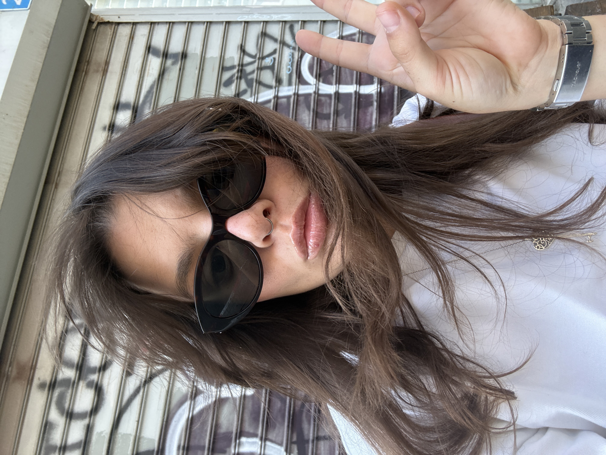 A young woman with shoulder-length brown hair, wearing black sunglasses and a nose ring, is making a peace sign with her hand. She is wearing a white shirt and a metal wristwatch. The background features a metal security gate with graffiti and possible structural elements of a building. The location is Tel-Aviv, Israel.
