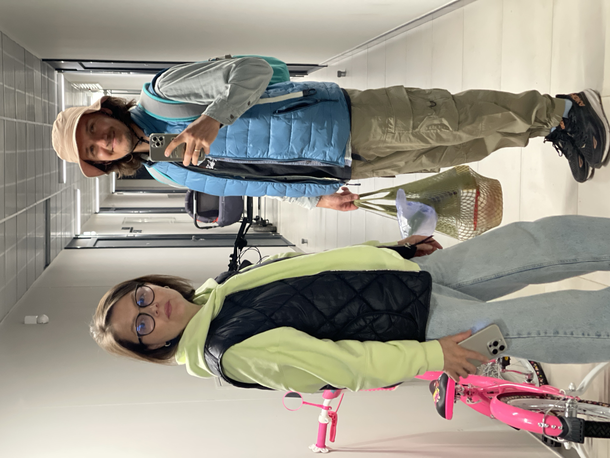 In an indoor hallway, a man and woman are present. The man, wearing a tan bucket hat, blue vest, grey shirt, and khaki pants, holds a shopping bag in his left hand and a smartphone in his right, suggesting he might be taking a selfie. A backpack is visible over his left shoulder. The woman, sporting glasses, a black vest over a neon yellow hoodie, and blue jeans, is seated on a pink children's bicycle. She also holds a smartphone in her right hand. The hallway is brightly lit with fluorescent lighting, featuring doors and a tiled wall on the left side. The location within the city is unspecified.