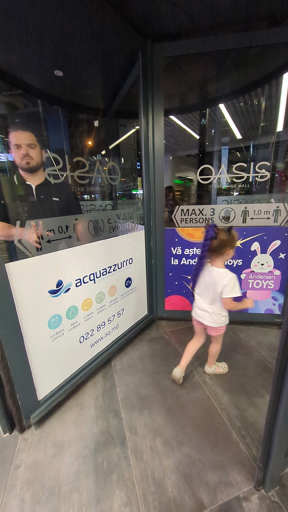 The image shows the entrance to the Oasis Shopping Mall in Chișinău, Moldova. A man is standing inside the entrance, reflected in the glass doors. A young girl is walking away from the camera, towards the right side of the image. The doors have signs indicating "MAX. 3 PERSONS" and "1.0 m" with figures of people, likely a social distancing sign. There is also a sign for "acquazzurro" which appears to be a dry cleaning service, with the phone number 022 89 57 57 and website www.aq.md. A colorful advertisement for "Andersen TOYS" is visible on the right side of the door. The floor is tiled.