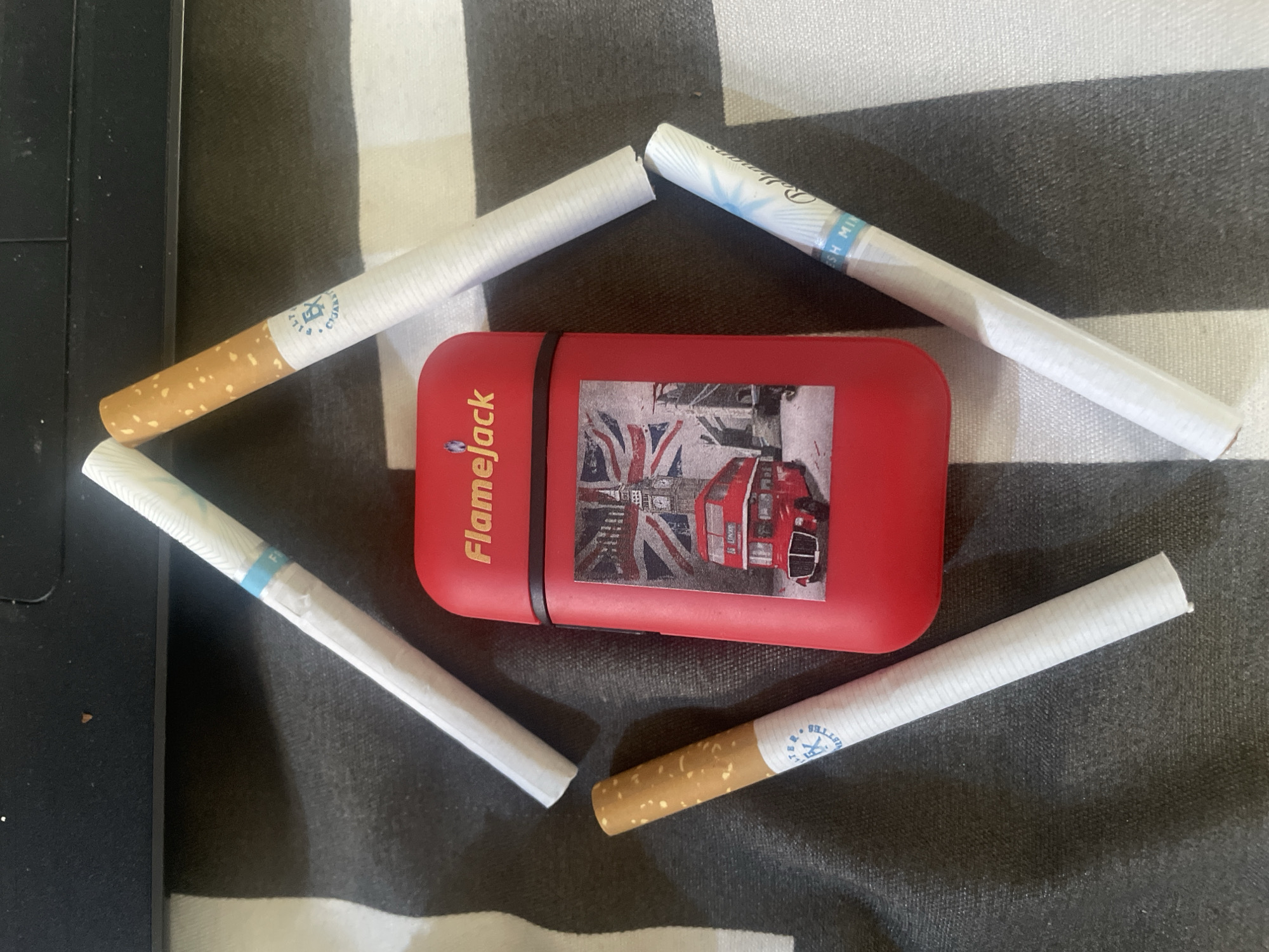 The image displays a red rectangular lighter with a London-themed decal, featuring Big Ben and a red double-decker bus, positioned centrally on a black and white striped surface. Four cigarettes are arranged diagonally around the lighter, forming a diamond shape. Two of the cigarettes have brown tobacco filters, while the other two have white filters with blue accents. One cigarette has text that reads "FILY EX CIGARETTES" and another has text that appears to include "SH MIN". The lighting suggests an indoor environment, possibly a desk or table, given the reflection on the black surface at the top of the image. There are no people or animals visible in the scene. The image does not contain any visual information that can be used to determine its location in Jos, Nigeria.