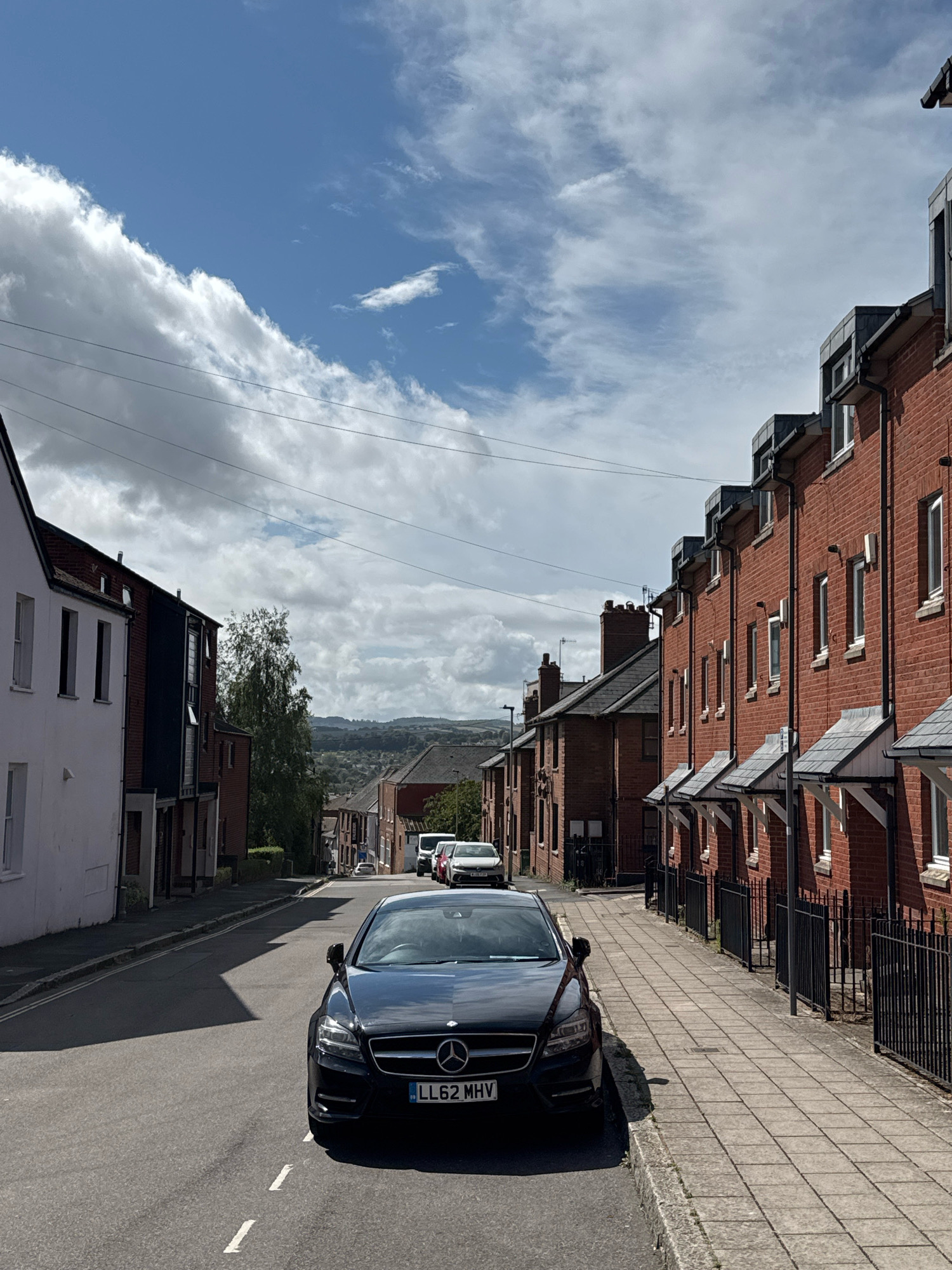A black Mercedes-Benz CLS 350 CDI, license plate LL62 MHV, is parked on the right side of a street in Exeter, United Kingdom. The car is facing away from the viewer and is positioned on the asphalt road, partially casting a shadow. To the right of the road, a paved sidewalk runs alongside a row of terraced houses with red brick facades and grey slate roofs. The houses have multiple windows and some have external lighting fixtures. A black metal fence separates the sidewalk from the properties. On the left side of the street, a white building with dark-framed windows is visible, followed by a building with a dark blue facade. Further down the street, more houses and trees can be seen, with rolling hills in the background under a cloudy sky with patches of blue. Several other vehicles are parked further down the street on the right. The scene appears to be a residential area.