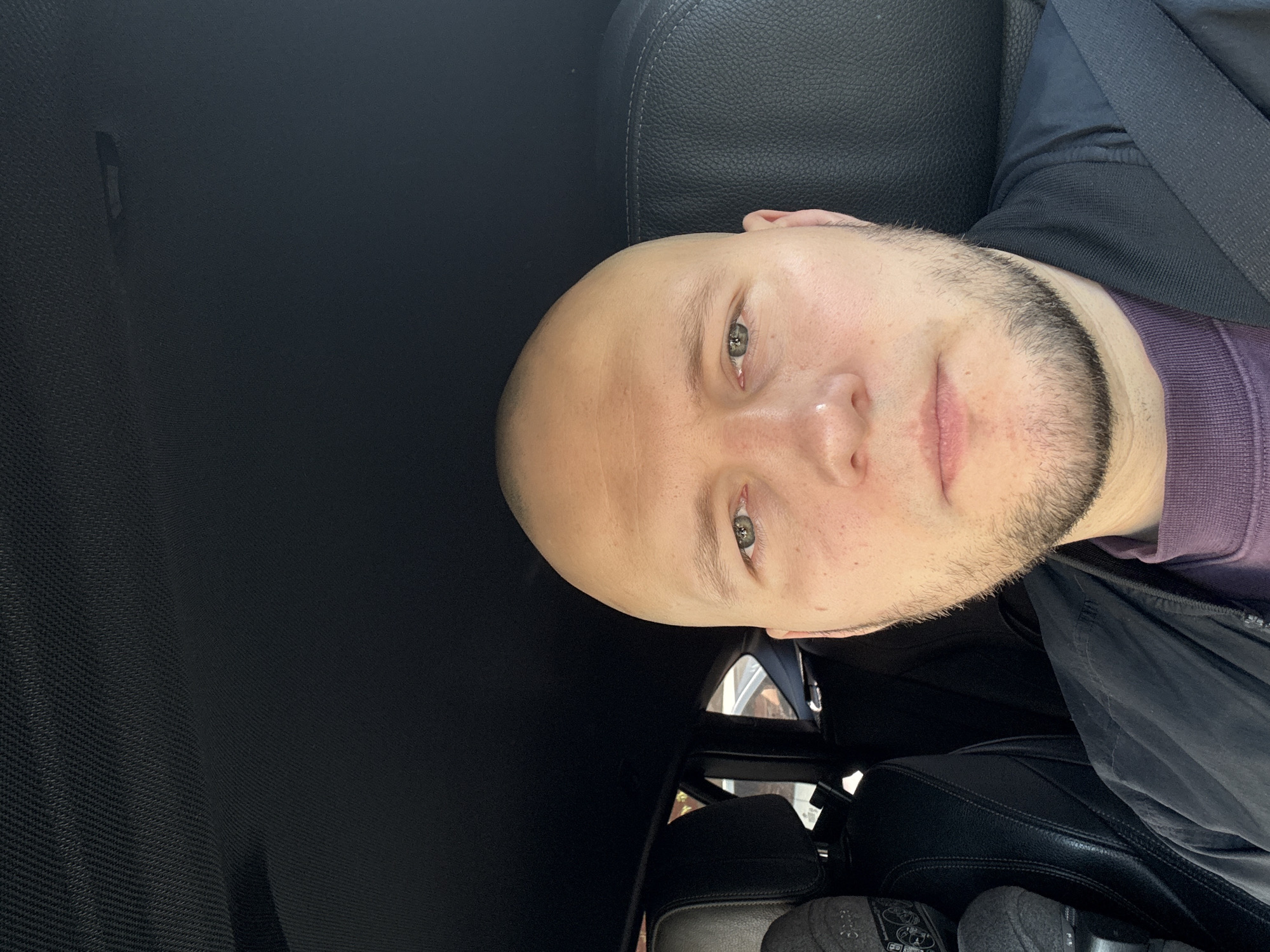 A bald man with a beard is sitting in a car. He is wearing a purple sweater and a black jacket. The car has black leather seats and a black interior. The location is unknown, but it cannot be confirmed to be in the city of Krasnoyarsk, Russia from the image alone.