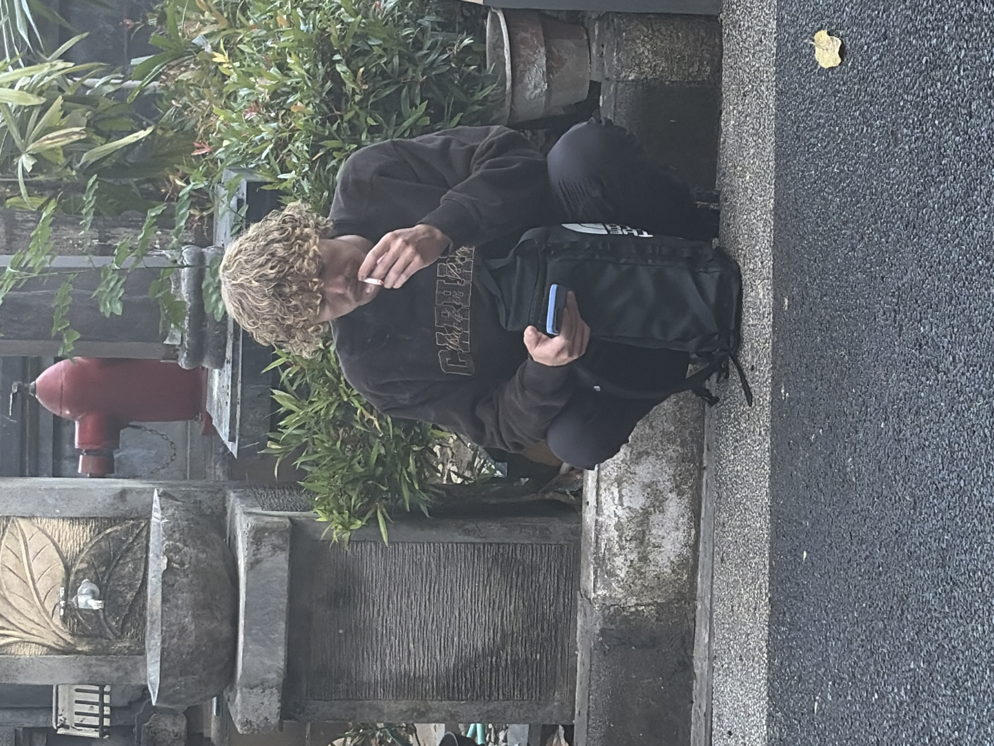 In Kuta Selatan, Indonesia, a young man with curly blonde hair is crouched down, smoking a cigarette and looking at his phone. He is wearing a dark brown sweatshirt and black pants. A black backpack is positioned in front of him. Behind him, a red fire hydrant stands against a stone structure adorned with a carved leaf motif and a faucet. Green foliage and a planter are visible to the right of the man. The scene takes place on a paved area with a bordering stone wall and walkway.