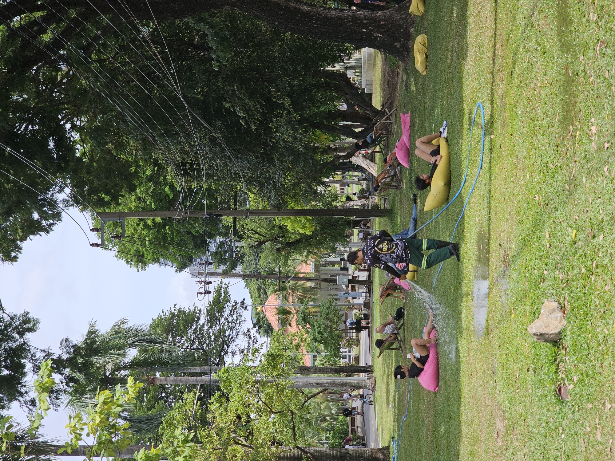 In Lumpini Park, Bangkok, Thailand, a man in green pants and a black shirt with a yellow logo waters a pink beanbag chair with a hose, creating a spray of water. A young girl in a black shirt and white shorts is seated on the beanbag, looking to her left. Nearby, a yellow beanbag chair is occupied by a young man in black shorts and a black t-shirt, lying down. Several other people are scattered across the green lawn, some sitting on chairs and beanbag chairs. A pathway is visible in the background, with people walking and a bicycle. Trees provide shade, and a building with a red roof can be seen further in the distance. Power lines crisscross the sky.
