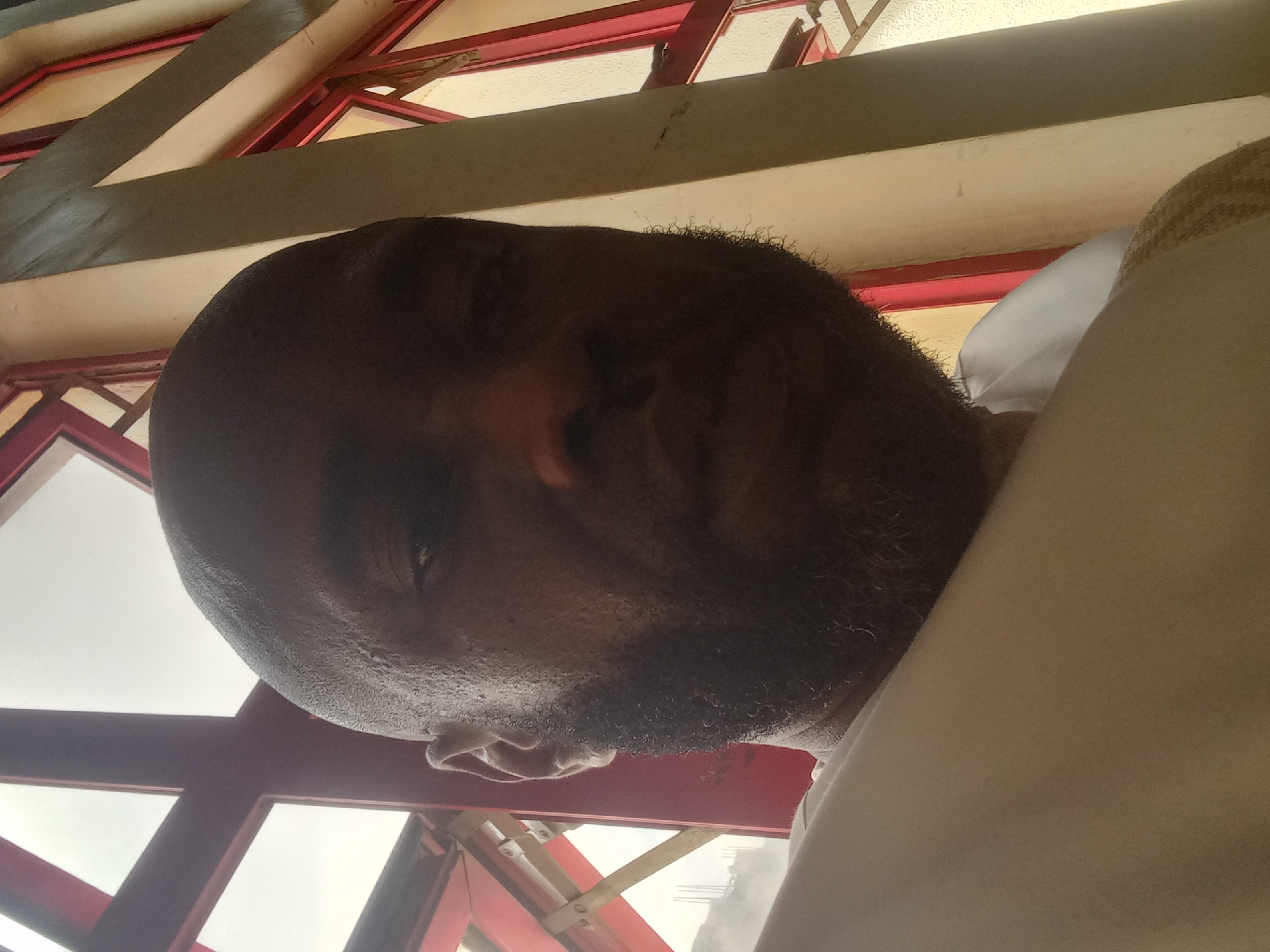The image is a close-up selfie of a bald, bearded man with dark skin. He is wearing a light-colored garment, possibly a shirt or robe. The background consists of a window frame with red and white painted elements, suggesting a building interior. The window reveals an exterior view, though details are unclear. The location is described as the city of Barden Yana, Nigeria. No specific actions or interactions are apparent.