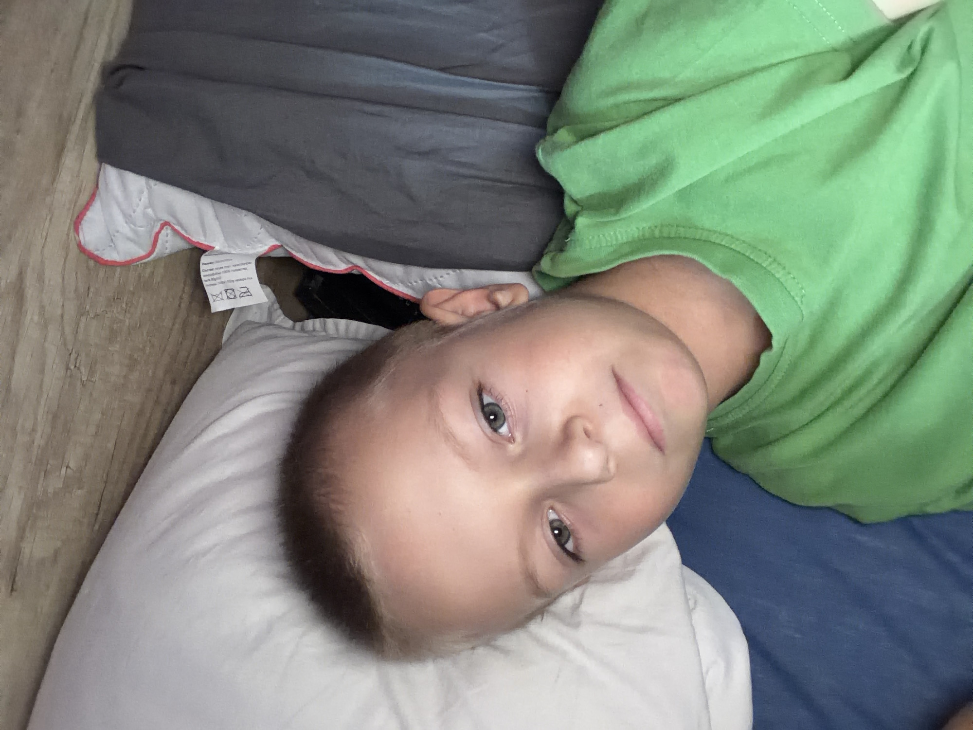 The image shows a close-up, high-angle shot of a young boy lying down. He is wearing a bright green t-shirt. His head is resting on a white pillow. Behind him, there is a wooden headboard and a gray blanket with a pink trim. A label is attached to the blanket. The setting appears to be a bedroom. The boy's eyes are open and he is looking towards the camera.