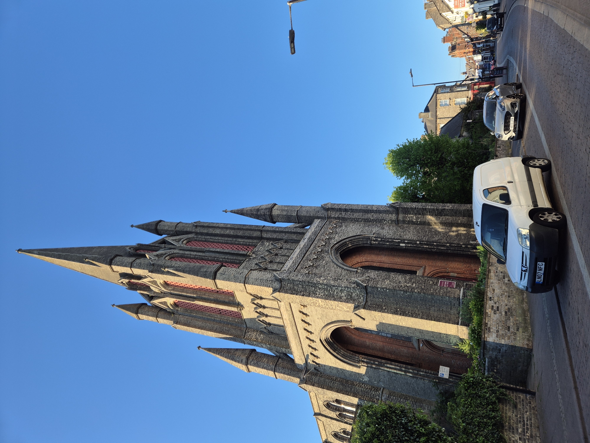 This image captures a prominent church building on a clear, sunny day in West Suffolk, United Kingdom. The photograph is taken from a low angle, looking upwards towards the church's impressive Gothic Revival architecture, characterized by tall spires, pointed arches, and a blend of grey and reddish brickwork. The warm, golden light and long shadows suggest it is late afternoon or early evening.
To the right of the church, a paved street is visible with two parked vehicles. In the foreground, there is a white Peugeot van with the license plate `LN07 NFC`. Further down the street, a grey Ford Puma is parked, displaying the license plate `LC21 YJF`. Beyond the street and cars, traditional British brick buildings line the road, and a "FOR SALE" sign can be faintly seen on one of them. A street light with a modern fixture extends into the bright blue sky from the right side of the frame. Lush green trees and bushes are scattered along the street and around the base of the church.
Notable details on the church include arched wooden doors and intricate stone and brick patterns. A small sign with a white 'P' (likely indicating parking) is visible on the church wall, and another sign with some red text is partially visible near one of the doors, though the full text is obscured. The scene depicts a quiet urban environment centered around a historic architectural landmark under a vibrant, clear sky.