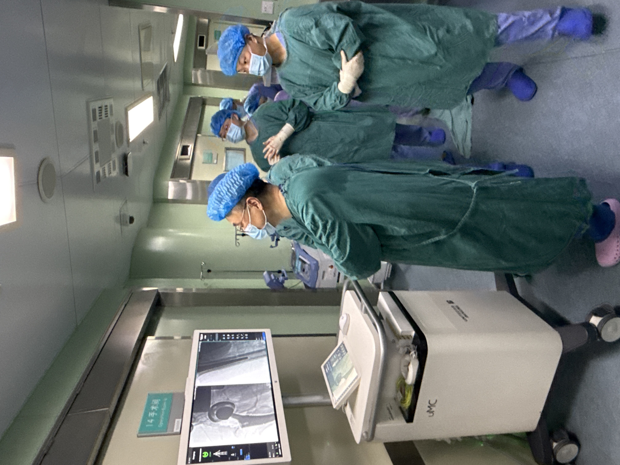 Inside a brightly lit operating room, likely located in a hospital in the Yuzhong District, China, four medical professionals, clad in green surgical gowns, blue surgical caps, and masks, are observing medical imaging displayed on a monitor. A mobile medical imaging unit with a screen and controls is stationed in the foreground. The monitor shows black-and-white images, likely X-rays, of what appears to be a prosthetic hip joint. A sign in Chinese script is visible on the wall next to the monitor.