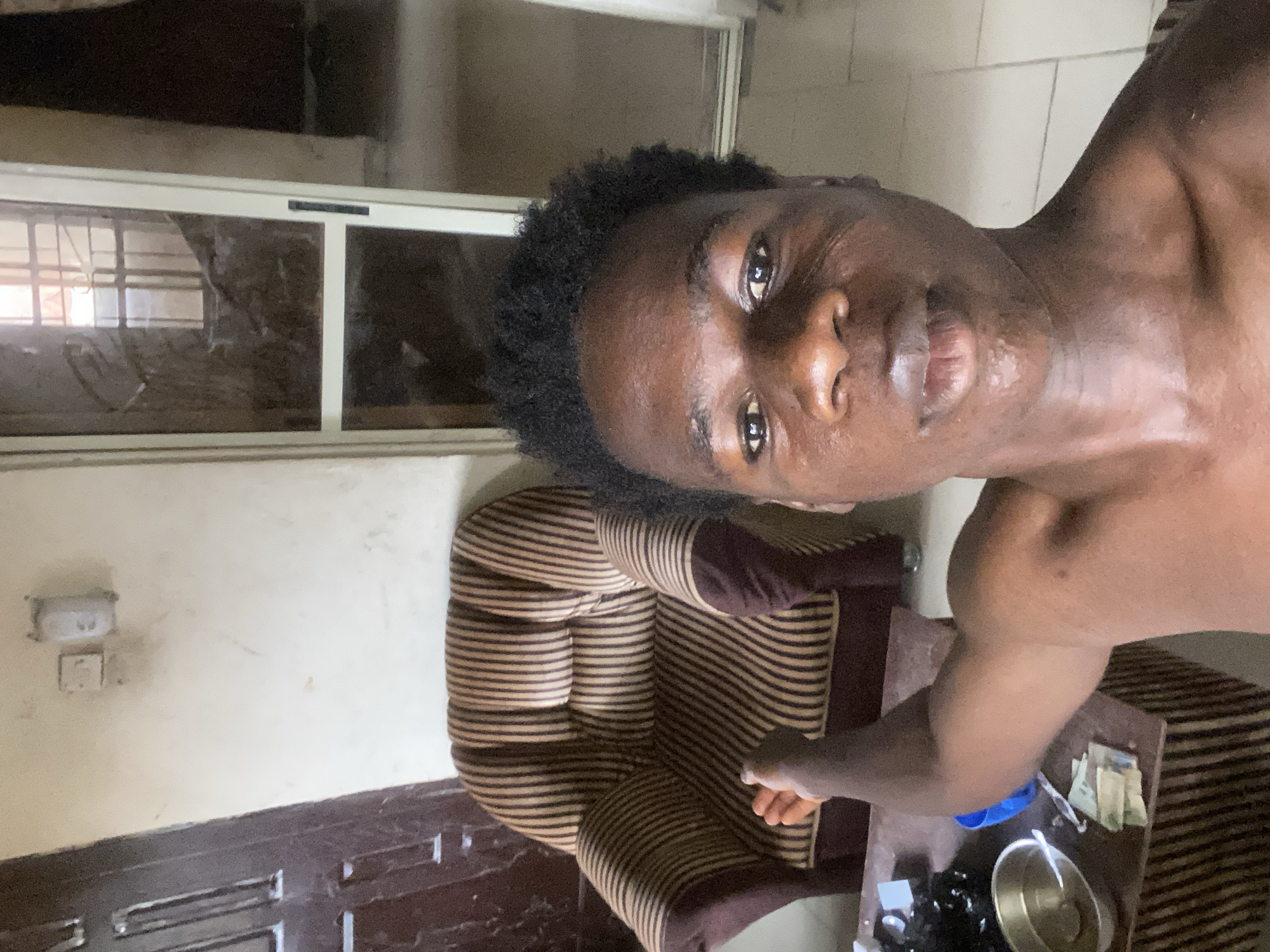 A shirtless, dark-skinned man is in a room in Mararaba, Nigeria. He has short, curly, dark hair. He is standing next to a striped brown and beige armchair. A dark wooden table is next to the armchair, holding a metal bowl, a blue container, and some paper currency. A white window frame is visible in the background. The room has white walls and a dark wooden floor with a patterned design. A light switch is on the wall.