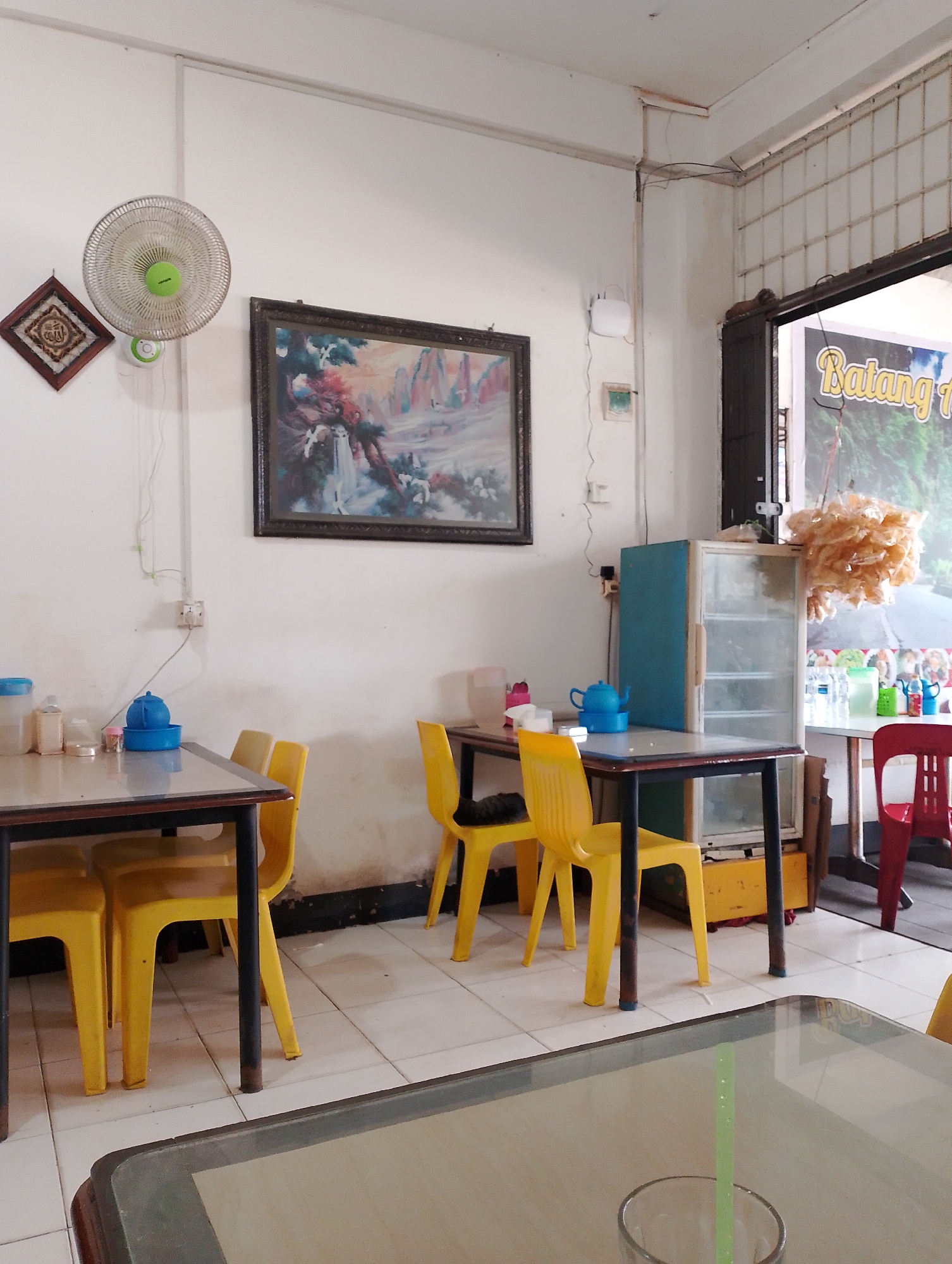 The image depicts the interior of a modest eatery, likely a warung or small restaurant, in Batam, Indonesia. The scene is set in a simple room with white walls and tiled flooring.
Dominating the left side of the image is a wall-mounted oscillating fan with a green center. To its left hangs a small, square decorative item framed in dark wood, featuring what appears to be Arabic calligraphy. Above and slightly to the right of the fan hangs a large, framed landscape painting with vibrant colors, depicting a scene with figures and possibly a waterfall.
In the foreground, two tables with dark frames and dark-colored table tops are visible. The table on the left has several items on it, including a blue teapot, a clear pitcher, and some plastic containers. Four bright yellow plastic chairs are arranged around this table.
To the right of the first table, there is a small, light blue and white cabinet with glass doors, displaying several shelves. Next to it is another table, partially visible, with a red chair pulled up to it. This second table has bottles of drinks and other items on its surface.
A large sign with yellow lettering, reading "Batang..." is visible through an open doorway on the far right, suggesting the eatery is located in Batam. Hanging from the ceiling near the doorway is a cluster of what appears to be dried food products, possibly shrimp crackers.
The overall impression is of a functional, no-frills establishment, likely serving local cuisine. The lighting is bright, suggesting daytime. There are no people visible in the image, and the scene is static. The environment appears clean but worn, typical of a local eatery.