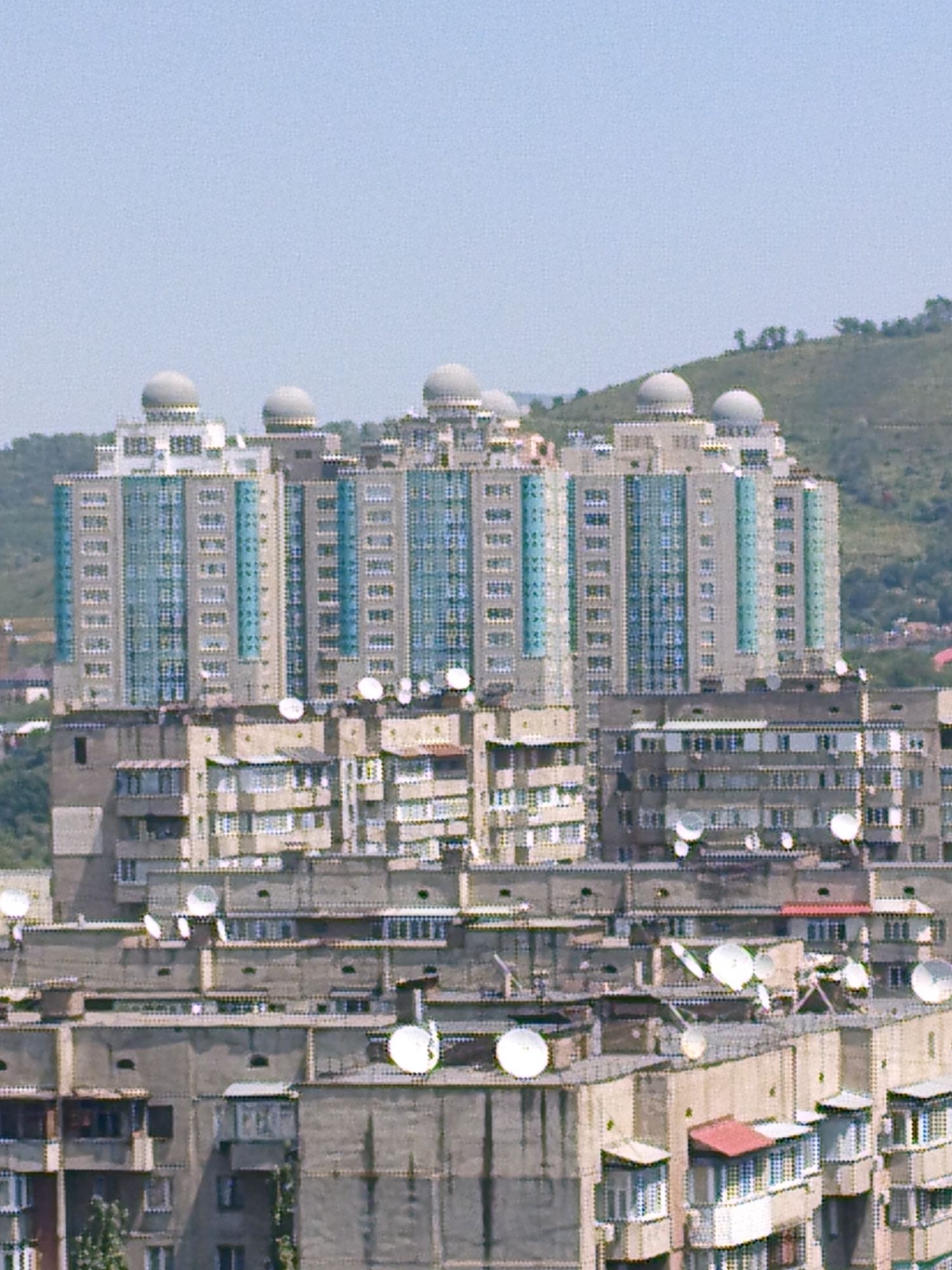 The image shows a cluster of apartment buildings in Almaty, Kazakhstan, with a residential building in the foreground and taller, modern buildings in the background. The foreground building is gray and appears to be an older Soviet-era construction with multiple balconies and satellite dishes on its roof. The background features several multi-story apartment buildings with blue accents and rounded domes on top, likely representing a more recent development. A green, hilly landscape is visible behind the buildings, suggesting an urban environment situated near a natural terrain. The sky is clear and pale blue. No people are visible in the image. The lighting suggests it is daytime.