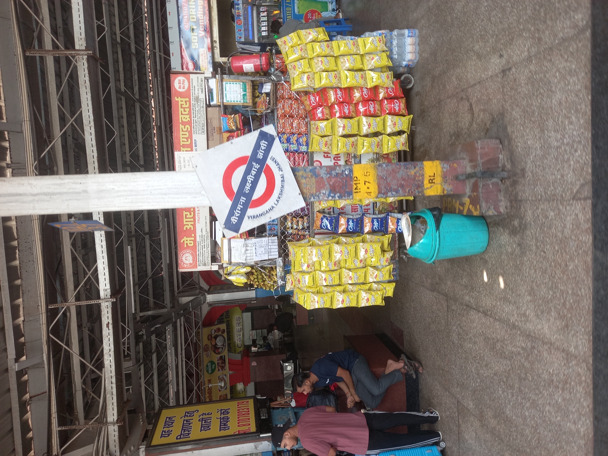 This image captures a scene at the Virangana Lakshmibai Jhansi Railway Station in Jhansi, India. A vendor's stall is visible, stocked with snacks like chips and water bottles. A blue trash can sits nearby. Two people are lying on the ground near the stall; one is wearing a maroon shirt and black pants, while the other is wearing a blue shirt and grey pants. Signage includes advertisements, information about food vendors, and the railway station name in Hindi and English. The station's infrastructure, including metal beams, is visible above the stall.