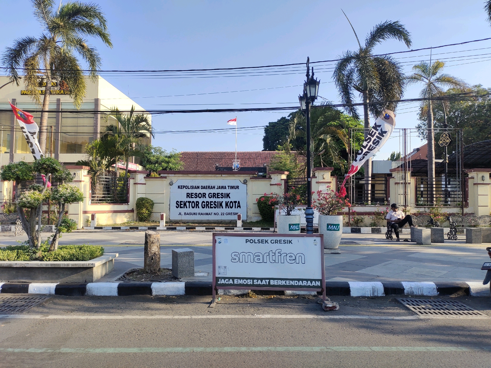 The image shows the front of a police station in Kebungson, Indonesia. The building is a light yellow color with large windows and a red-tiled roof. In front of the building, there is a sign that reads "KEPOLISIAN DAERAH JAWA TIMUR RESOR GRESIK SEKTOR GRESIK KOTA JL. BASUKI RAHMAT NO. 22 GRESIK". To the left of the sign, there is a flag with the Indonesian colors of red and white. There are also several palm trees and a street lamp. In the foreground, there is a Smartfren advertisement sign. A person is sitting on a bench in front of the building, and there are several other benches visible. The sky is clear and blue.