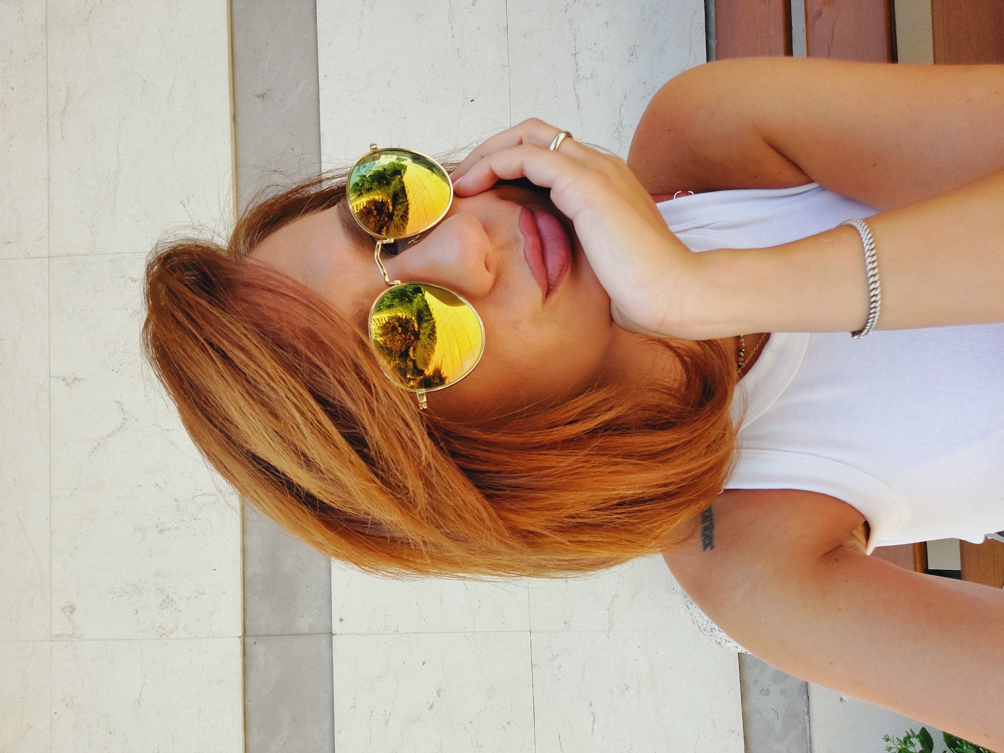 A woman with auburn hair, wearing a white tank top and round, gold-rimmed sunglasses with yellow lenses, rests her chin on her hand. Her lips are slightly pursed. A silver bracelet adorns her wrist, and a ring is visible on her ring finger. A tattoo of Roman numerals, "XXII", is visible on her left shoulder. She is seated on a wooden bench against a tiled wall. The sunglasses reflect a sunny outdoor scene with trees and a path. This appears to be an outdoor setting in Sofia, Bulgaria, likely a public space such as a park or a café with outdoor seating.