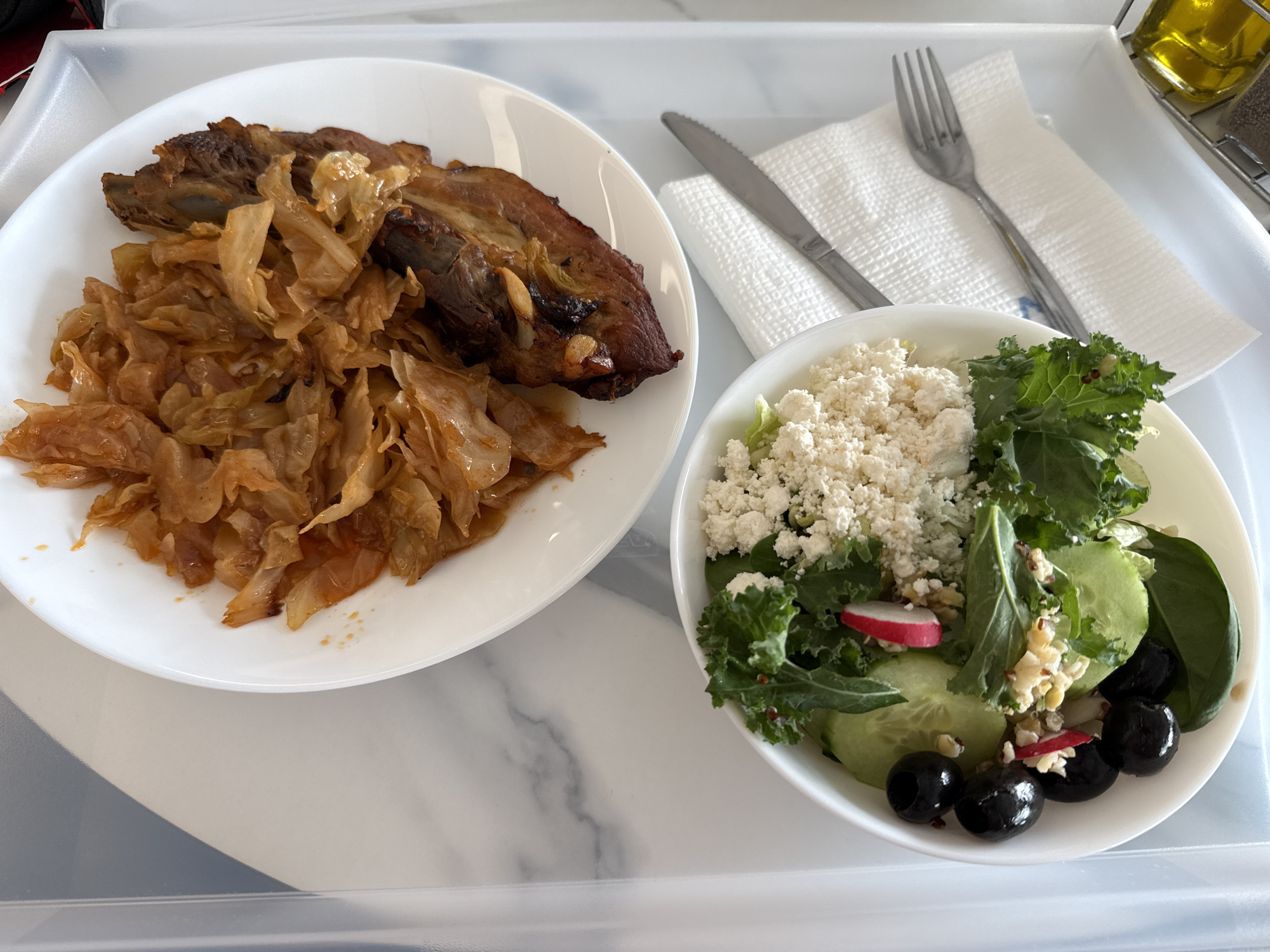 The image presents a meal on a white tray. A white plate on the left holds cooked cabbage and a piece of roasted meat, possibly pork. To the right, a white bowl contains a salad with fresh greens, cucumber slices, radishes, black olives, and crumbled white cheese. A knife and fork rest on a white napkin beside the bowl. A bottle of liquid, likely olive oil, is visible in the upper right corner. The tray has a white marble-patterned surface. The scene appears to be indoors, potentially on a flight or in a hospital setting, based on the tray presentation. There is no information in the image to suggest it is taken in Sofia, Bulgaria.