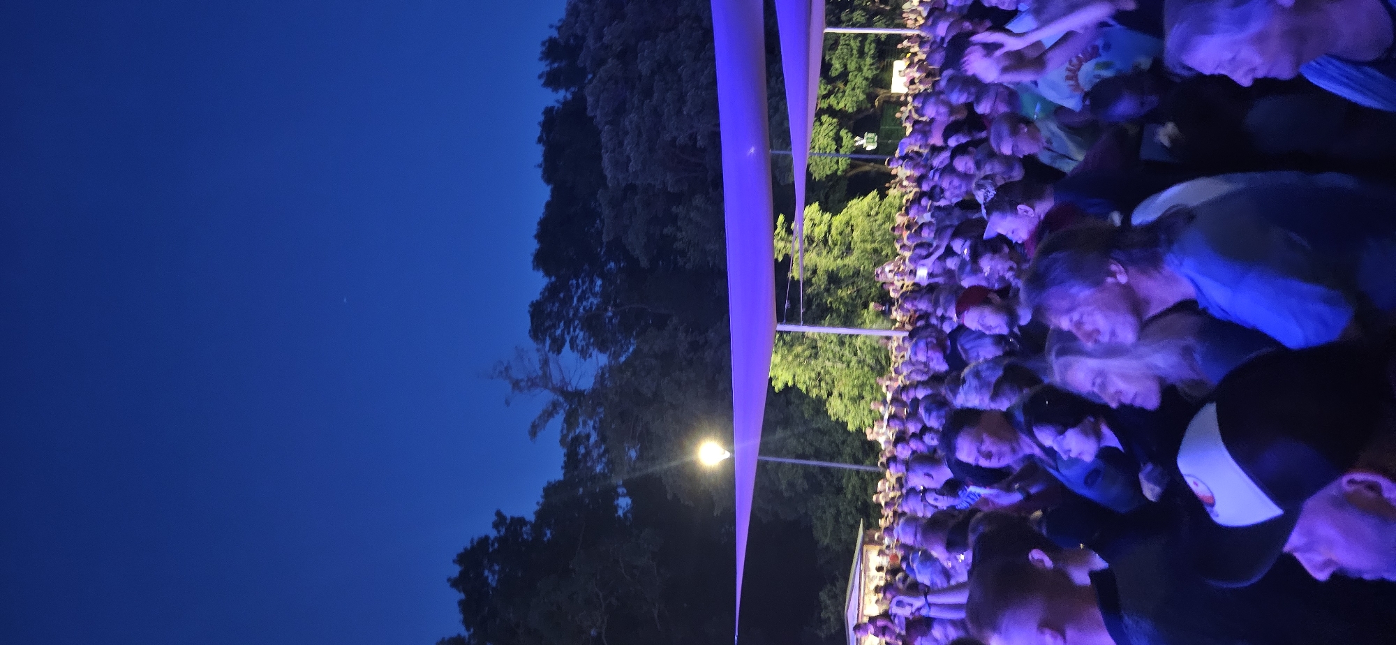This media file depicts an outdoor gathering of people at dusk, with a deep blue sky suggesting it is twilight. The scene is illuminated by a bright, warm light source that appears to be a street lamp, and a vibrant purple structure, likely a temporary awning or stage covering, stretches across the frame, casting a colored glow on the crowd.
The subjects are a large crowd of individuals, mostly facing away from the camera and towards an unseen focal point, suggesting they are attending an event such as a concert or festival. The people are a mix of ages and genders, dressed in casual attire. The overall emotion conveyed is one of engagement and shared experience, with many looking intently towards the event.
Notable details include the lush green trees visible behind the crowd, hinting at a park or outdoor venue setting. The presence of a light pole and the purple structure suggest a prepared event space. The time of day is clearly evening, given the sky color and artificial lighting. While the location is Hévíz, Hungary, there are no specific visual cues within the image itself to confirm this. No visible text is present in the media file.