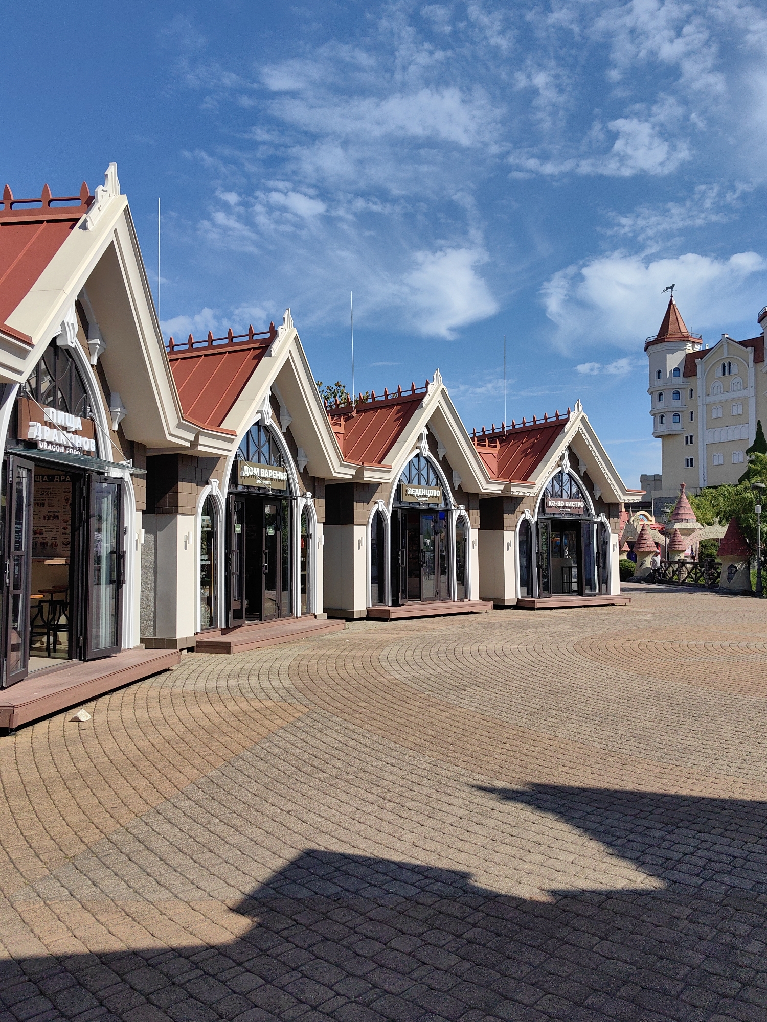 The image shows a sunny day in Sirius, Russia, with a clear blue sky and scattered white clouds. In the foreground, a paved courtyard with a circular pattern of bricks leads to a row of shops with red roofs and arched windows. Signs above the entrances indicate various establishments, including "Dom Varenya" (House of Dumplings) and "Pizza Drakon" (Dragon Pizza). To the right, a large, light-colored building with a tall tower and red roof, resembling a castle, stands against the sky. The courtyard is partially shaded by the buildings. In the background, more whimsical, castle-like structures and green trees are visible.