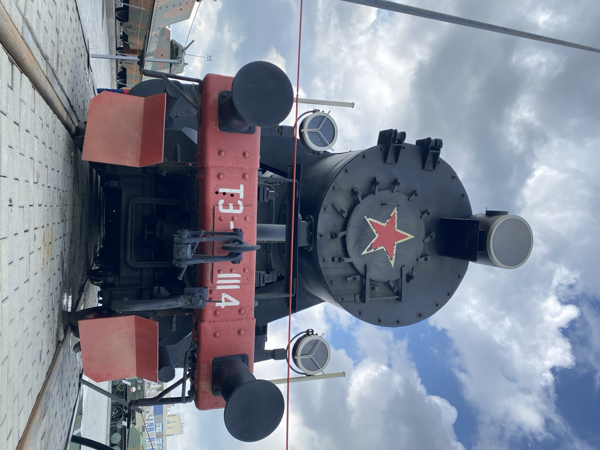 An outdoor, low-angle, eye-level shot captures the front of a black steam locomotive, identified by the Russian inscription "T3-1114" on its red bumper. The locomotive is adorned with a prominent red star emblem. To the left and right of the locomotive's front, two round, glass-paned lights are visible, mounted on tall, thin poles. The scene also includes several armored vehicles in the background, suggesting a museum or historical display. The sky above is partly cloudy with a mix of blue sky and white clouds. The locomotive is situated on railway tracks made of concrete slabs.