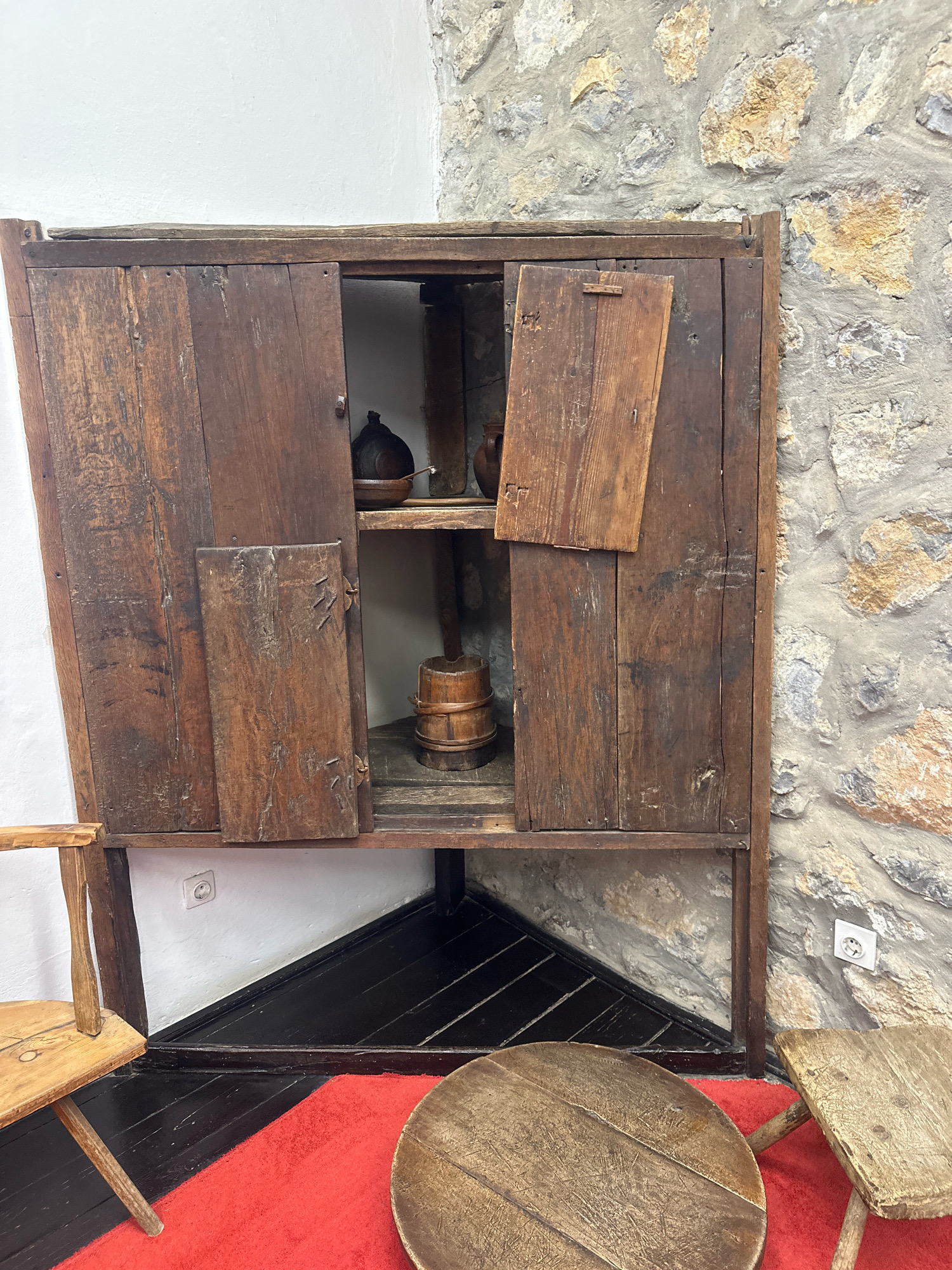 This is a medium-full shot of an antique wooden cabinet with its doors open, displayed against a stone wall. The cabinet, appearing to be made of rough-hewn wood planks, has two doors on the left side and one on the right, all partially open. Inside the cabinet, on shelves, are various antique items. On the top shelf, to the left, is a dark, possibly ceramic or metal object with a rounded top, resembling a jug or pot. To its right is a wooden bowl. On the lower shelf, a small, wooden barrel with metal bands is visible. The cabinet stands in a corner, with a black-painted floor and a red rug in the foreground. To the left of the cabinet, a rustic wooden chair is partially visible. To the right, a wooden stool with a triangular seat is also partially visible. The setting suggests an indoor display, possibly a museum or an antique shop. The location is identified as Topola Municipality, Serbia. The overall scene conveys a sense of history and tradition through the aged furniture and rustic decor.