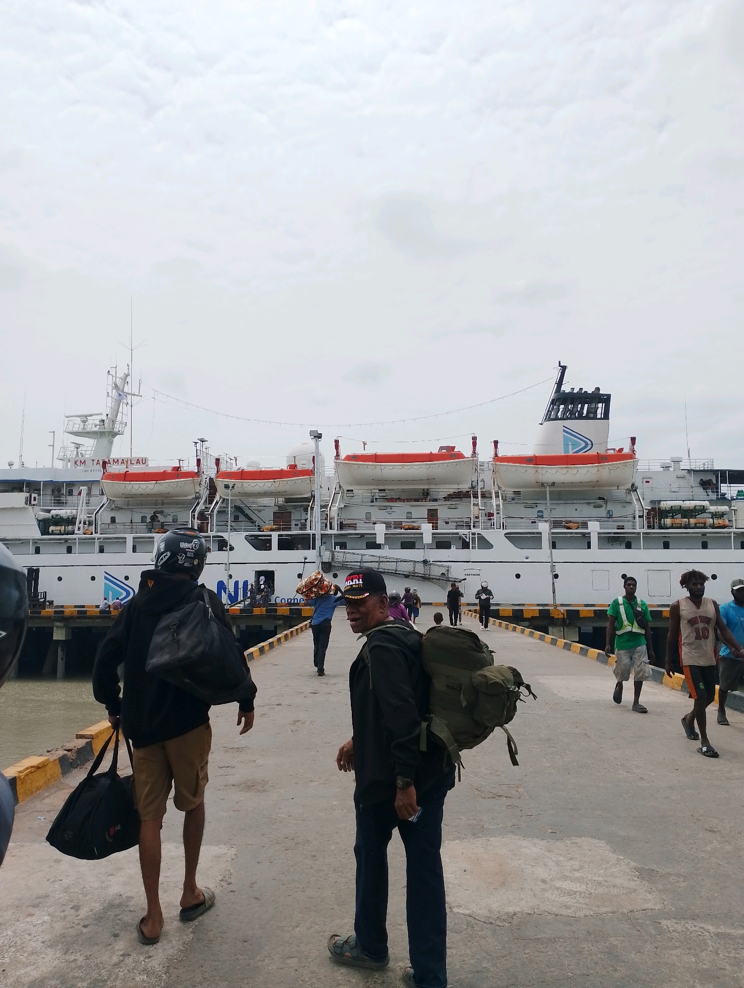 The image depicts a scene at the Merauke Port in Merauke, Indonesia, on a cloudy day. A large white ferry, the KM. TATAMAILAU, is docked alongside a pier. Several people are seen disembarking or walking on the pier. In the foreground, two men are walking away from the camera. The man on the left is wearing a black hoodie, khaki shorts, and sandals, carrying two black bags. The man on the right is wearing a black jacket and dark pants, with a large green backpack. Further down the pier, other individuals are walking, some carrying bags. Lifeboats are visible on the side of the ferry. The pier has a yellow and black striped railing. The water is visible on the left side of the image.