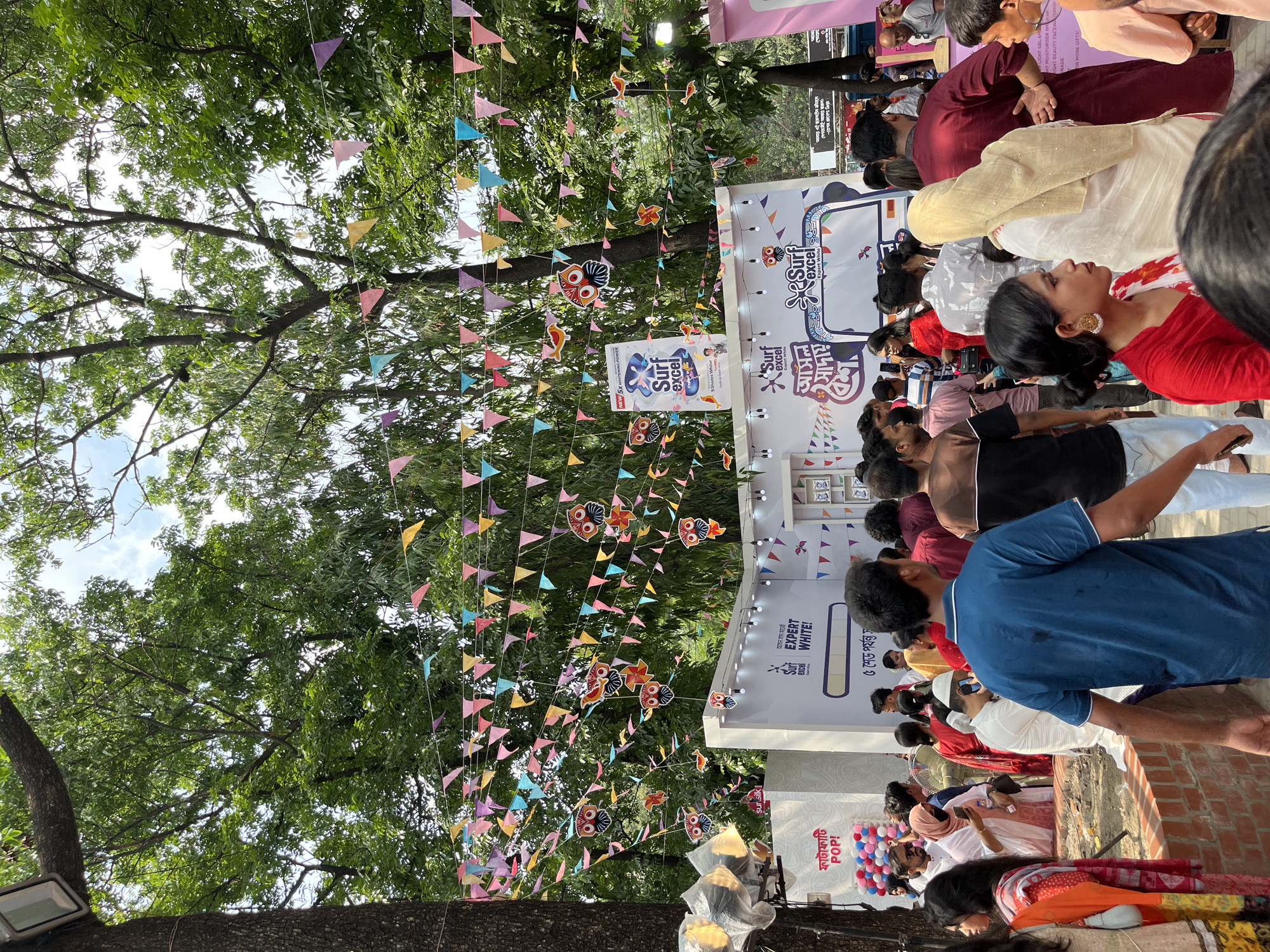 The image shows an outdoor event, possibly a promotional gathering, likely in Dhaka, Bangladesh. A crowd of people is gathered around a promotional booth for "Surf Excel," a laundry detergent. Banners and colorful pennants decorate the area, strung between trees. The scene is partially shaded by the tree canopy, indicating a daytime setting. The crowd includes people of various ages, wearing a mix of traditional and contemporary clothing. The booth displays "Surf Excel" advertisements. There are balloons visible near the bottom of the frame.