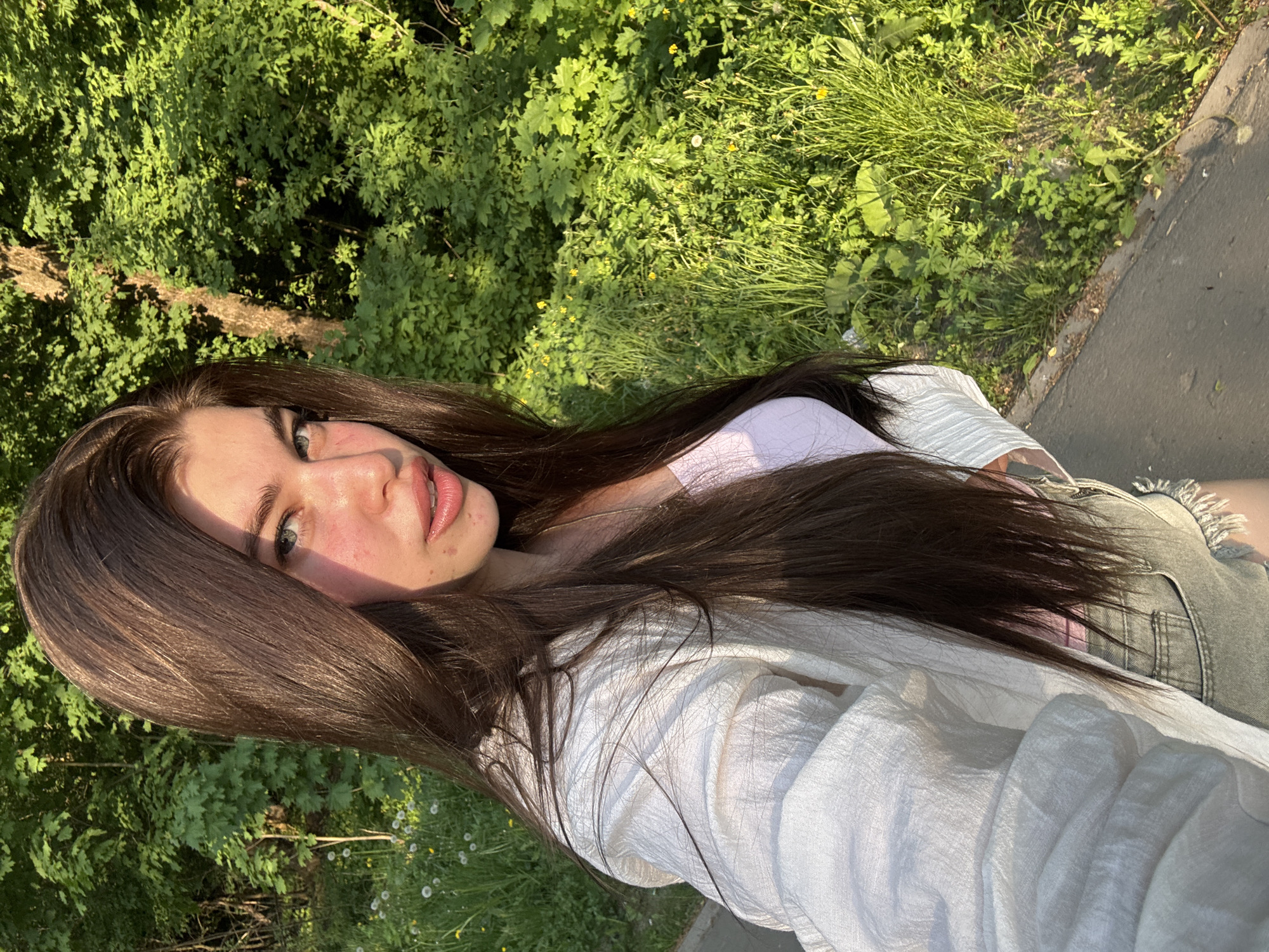 The image shows a woman with long, dark brown hair. She is wearing a white shirt and what appears to be a light purple top underneath. She is also wearing denim shorts. The background consists of green foliage. The location is possibly Посёлок комбината стройматериалов-2, Russia based on visual details like vegetation and architectural elements, though precise confirmation requires on-location information. The photo appears to be taken during daytime with natural lighting.