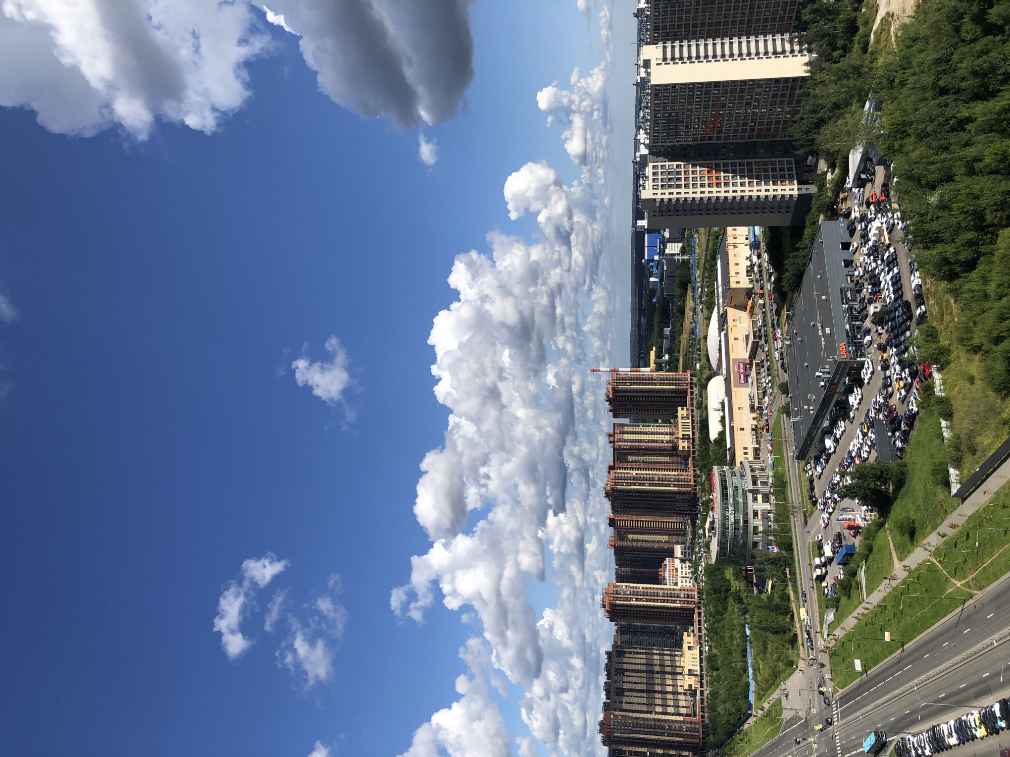 A high-angle, eye-level, full exterior shot captures a dense urban landscape under a bright blue sky dotted with puffy white clouds. The foreground is dominated by a multi-lane road with parked cars on the left side and a paved walkway. Beyond the road, a large parking lot is filled with numerous vehicles, a circular building with a glass facade, and a long, low-rise commercial building with a visible sign that reads "Dionisi".
Further into the scene, a row of identical, tall, modern apartment buildings with orange and brown facades stands prominently. To the right, another cluster of equally tall apartment buildings with cream-colored facades and orange window frames is visible. The background reveals a sprawling urban environment with more buildings of varying heights, some industrial structures, and a distant treeline. The overall scene suggests a contemporary, densely populated city area on a clear day.
