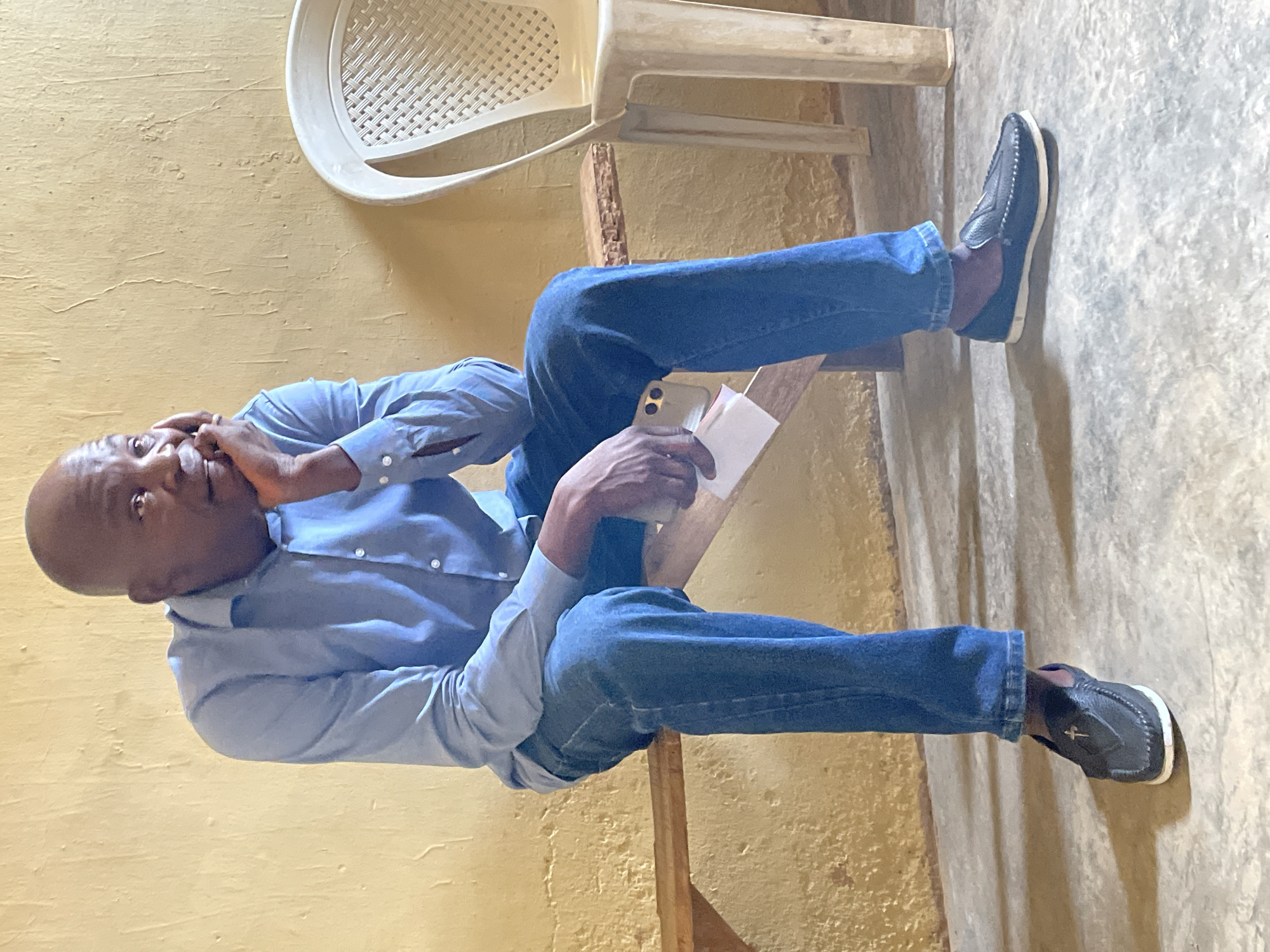 An adult man, bald and with dark skin, is seated indoors, looking slightly away from the camera with a pensive expression. He is wearing a light blue long-sleeved button-up shirt and blue jeans, complemented by dark moccasin-style shoes with white soles and a distinctive light-colored symbol on top. His right hand is raised, with fingers resting near his chin or cheek. In his left hand, he holds a smartphone, visible by its camera array on the back, along with a small white piece of paper or card.
He appears to be sitting on a simple wooden structure, possibly a bench or chair base. The background features a pale yellow textured wall. Above and behind him, a light-colored plastic chair with a woven back design is visible, positioned upside down or tilted. The floor is a light-colored, uneven concrete surface. The lighting is diffused, suggesting an indoor setting in Jos, Nigeria. There is no discernible text on the paper, phone, or shoes.