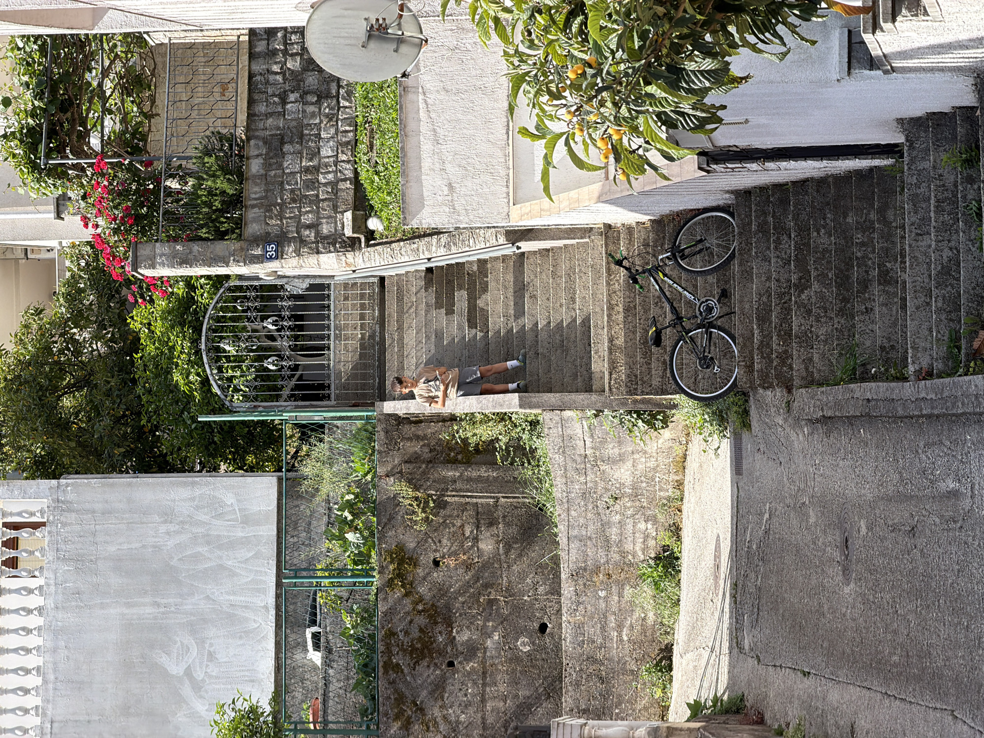 An eye-level, rotated aerial shot depicts a person lying on a set of outdoor concrete stairs in what appears to be an urban setting. The person is lying on their back, facing upward, wearing a light-colored shirt and shorts. A bicycle is positioned on the stairs above the person. The stairs lead from an upper level with buildings and vegetation down to a lower level. To the left of the stairs is a concrete wall with vegetation growing along it, and a wire fence with a vehicle visible behind it. Number 35 is on a small stone wall next to a gate near the stairs. The buildings appear to be residential and constructed of concrete.
