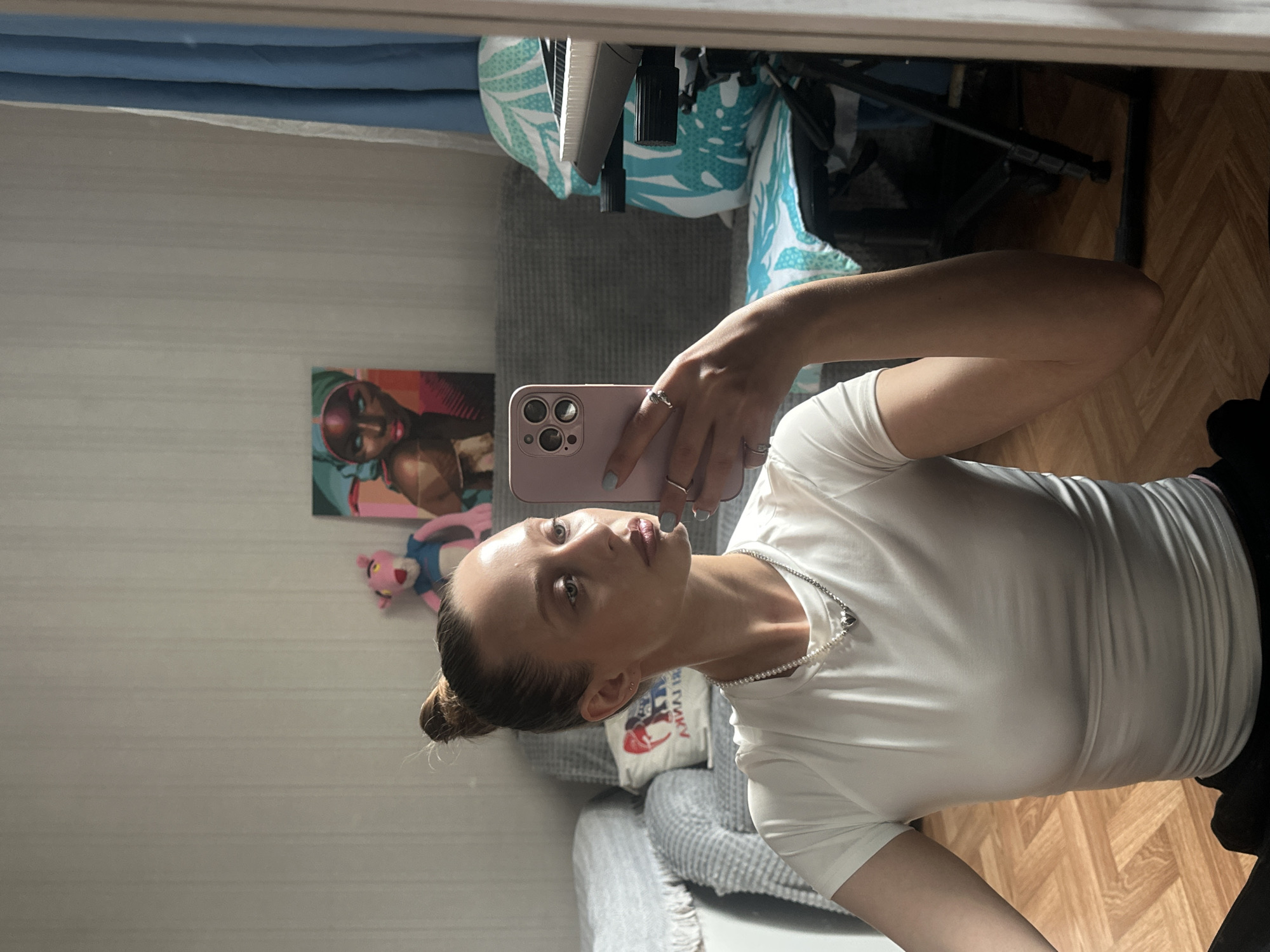 A young woman with blonde hair in a bun is taking a selfie in a room. She is wearing a white t-shirt and a silver necklace with a heart pendant. She is holding a pink phone in her right hand. In the background, there is a couch with pillows, a pink stuffed animal, a painting of a woman, and a keyboard. The room has light-colored walls and a wooden floor. The image appears to be taken indoors, possibly in an apartment in Khimki, Russia.