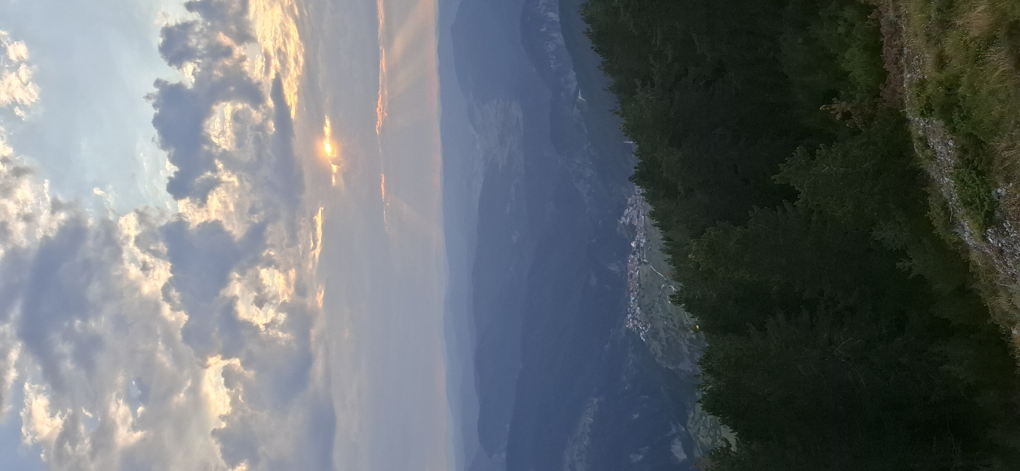 A vertical, eye-level, wide shot captures a serene view of the village of Gyovren, Bulgaria, nestled within a valley and framed by a dense forest of pine trees in the foreground. The sky above is dramatic with large, textured clouds, some tinged with the warm hues of sunrise or sunset, with rays of light piercing through. The sun itself is partially obscured by clouds, casting a soft glow. Layers of misty blue mountains recede into the distance, creating a sense of depth. The village is visible on a hillside, characterized by its red-tiled roofs and clustered buildings. The forest in the foreground consists of dark green pine trees, with a rocky and grassy embankment at the bottom of the frame. The overall atmosphere is tranquil and picturesque.