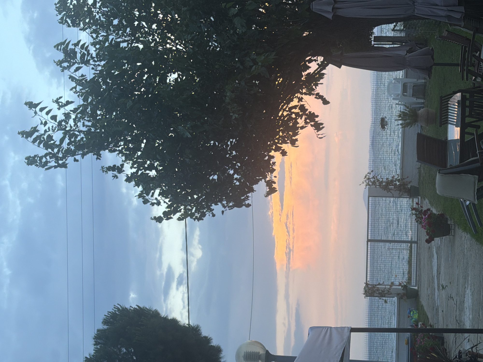 The image shows a serene outdoor scene at sunset, likely in Mandra, Greece. The sky is a canvas of soft blues and pinks, with clouds tinged with orange from the setting sun. In the foreground, a paved pathway leads to a grassy area with outdoor furniture, including a table with chairs and several closed umbrellas. Lush green trees frame the scene, their leaves casting dappled shadows. Beyond the paved area, a metal fence separates the property from the calm sea, which reflects the colors of the sky. A few potted plants and a decorative lamp post add to the ambiance. The overall atmosphere is peaceful and tranquil, suggesting a quiet evening by the sea.