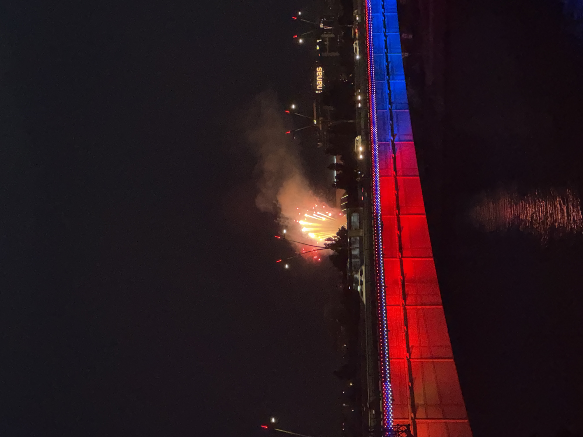 The image depicts a night scene at the Ada Bridge in Belgrade, Serbia, during a fireworks display. The bridge is illuminated with red and blue lights along its railings, creating a vibrant contrast with the dark sky. Fireworks are exploding in the upper right portion of the frame, casting a warm glow and generating plumes of smoke. In the background, city lights are visible, and a building with the illuminated sign "nanas" can be identified. Vehicles are seen moving on the bridge and on a road in the distance. The surface of a body of water, likely the Sava River, reflects the bridge's lights. There are no people clearly visible in the image. The overall atmosphere is festive and celebratory.