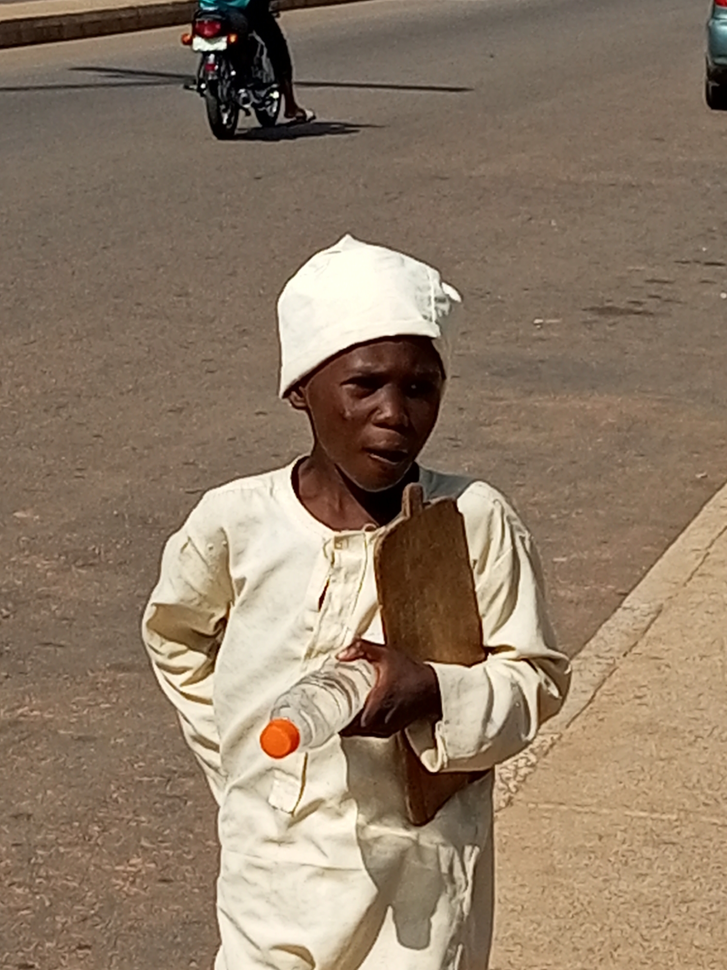 A young boy wearing a white skullcap and a cream-colored, long-sleeved tunic stands on a paved surface. He is holding a clear plastic water bottle with an orange cap in his right hand, and a wooden board in his left arm. The boy's mouth is slightly open, and he is looking towards the viewer. In the background, a motorcycle with a rider is visible on a road. The scene appears to be in the city of Tumfure, Nigeria, as indicated by the context. The lighting suggests it is daytime, with shadows cast on the road.