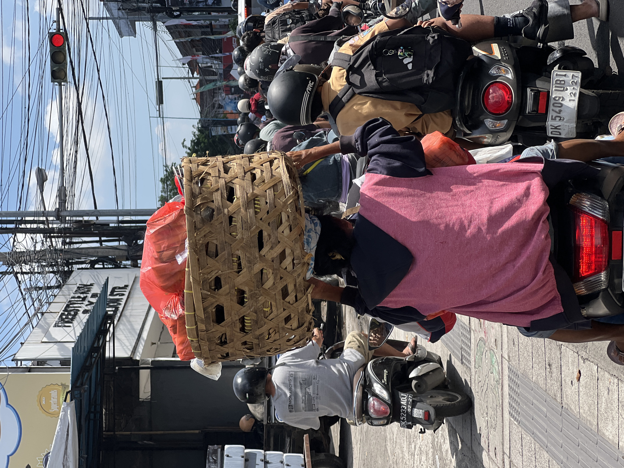 This image shows a congested street scene in Denpasar, Indonesia, with numerous motorcycles packed closely together. The vehicles are stopped at a red traffic light. In the foreground, a person on a motorcycle is transporting a large, woven basket filled with red bags and other items. Other riders are wearing helmets and carrying backpacks. The license plate on the motorcycle in the foreground reads "DK 5233 ADJ" with an expiration of "02-27." A sign for "FREESTYLE KON" on "LION STR." is visible above the street.