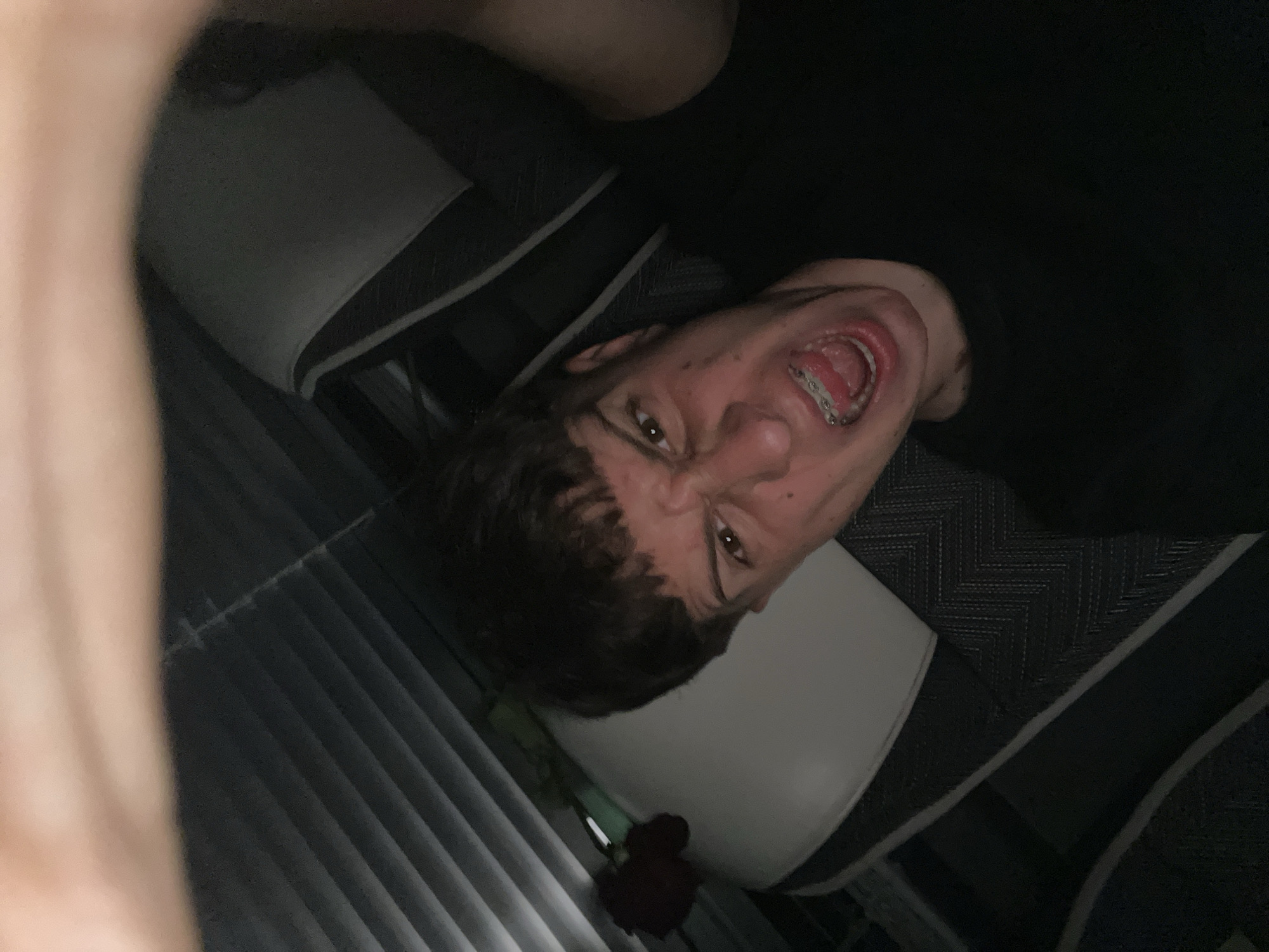 A low-angle photograph shows a person with short brown hair and visible dental braces, lying on what appears to be a patterned car seat. The person's mouth is open and their face is distorted in an apparent grimace. To the lower left of the person is a red flower laying on closed window blinds. The environment is dimly lit. There is no indication of the location being Saraia, Bulgaria.