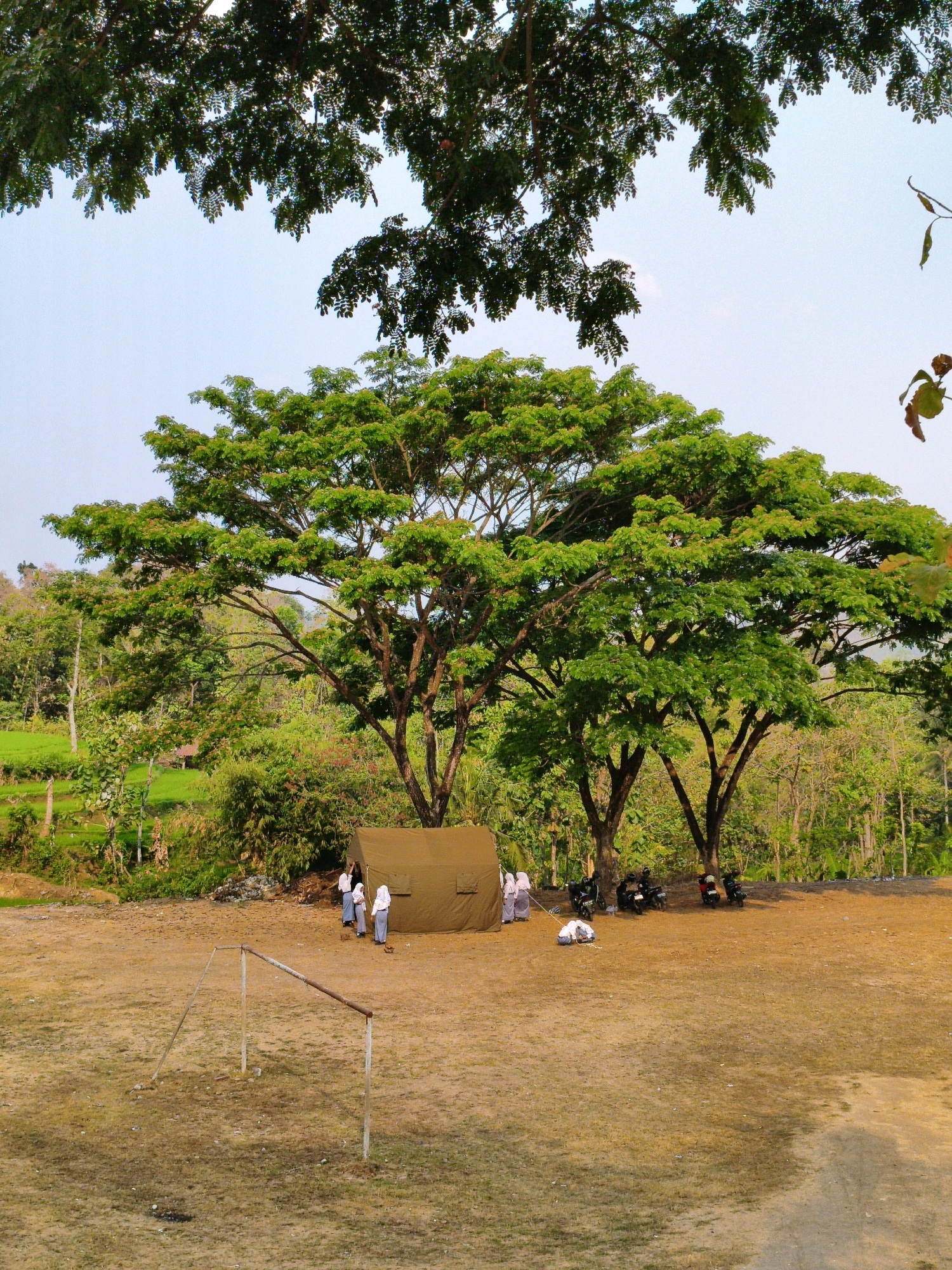 The image depicts an outdoor scene, likely a field, in Puhpelem, Indonesia. In the foreground, a rudimentary metal goalpost, weathered and rusted, stands on dry, patchy grass. Behind it, a large, khaki-colored tent is pitched, surrounded by several motorcycles parked in the shade of large, leafy trees. A group of three individuals in white and blue uniforms, appearing to be students, are standing near the tent. Two of them are looking at something on the ground near the tent, while another stands behind them. The background is filled with lush green vegetation, including trees and what appears to be a rice paddy field. The sky is a pale blue with hints of clouds. The overall impression is a rural setting, possibly a school ground or a campsite, with students engaged in an activity under the shade of trees.