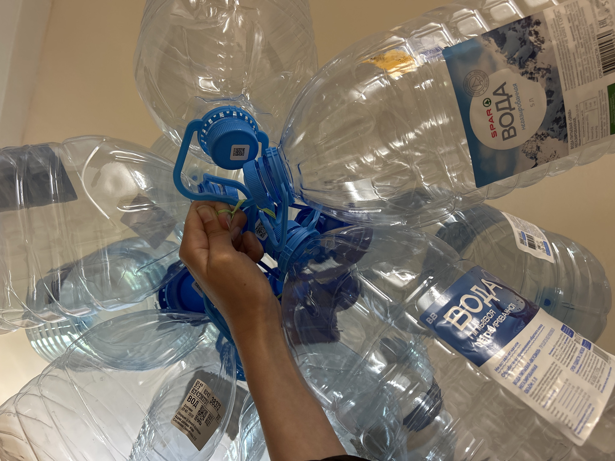 A cluster of empty, clear 5-liter plastic water bottles hangs from a light fixture in a room in an Unknown, Unknown location. Most of the bottles still have their blue caps and handles attached, and several have their original labels. The brands visible include "SPAR" and "ВКУСВИЛЛ". A person's arm and hand reach up to the cluster, their fingers apparently manipulating a green string or tie among the blue bottle handles. The bottles appear to be arranged in a vaguely floral or star-shaped pattern. The background is a plain off-white or light beige wall.