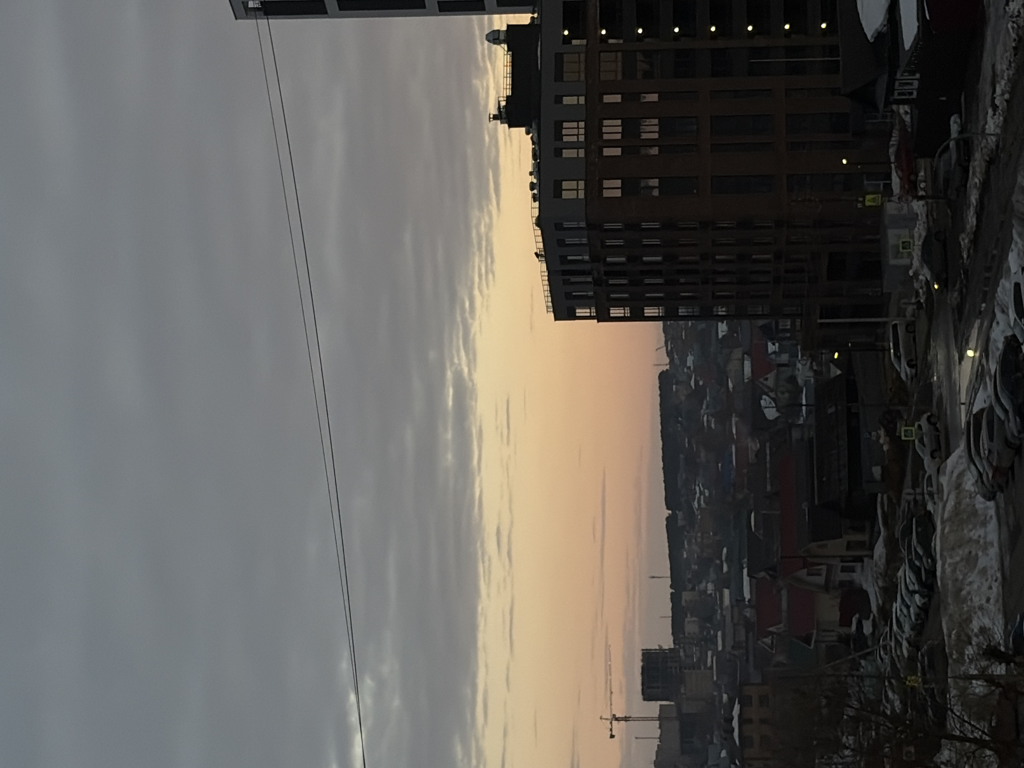 The image displays an urban landscape in Izhevsk, Russia, under low light conditions consistent with twilight or an overcast winter/early spring day. The sky features stratified grey clouds, with a faint band of orange-pink light visible along the distant horizon. Two dark utility lines stretch horizontally across the upper mid-sky.
In the foreground and midground, streets and parking areas are partially covered with dirty snow and slush, accommodating numerous parked vehicles, primarily sedans and SUVs. On the right side, a multi-story building with a dark, modern facade and illuminated windows rises prominently, with a taller, narrower building partially visible to its right. To the left and center, a mix of smaller, varied-roof residential and commercial structures are present. In the background, a large yellow tower crane and a building under construction are visible against the horizon line, which is topped by a dark, possibly forested, ridge.
No people or active interactions are discernible within the frame.