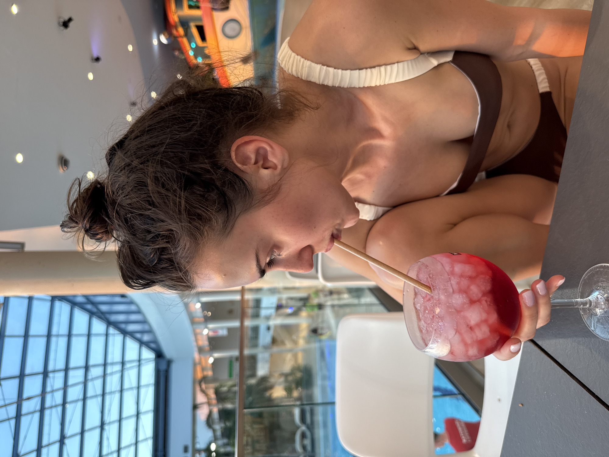 A woman in a brown and white bikini is drinking a red beverage with a straw in an indoor environment. The woman has brown hair pulled back in a bun, and her eyes are closed as she sips from the glass. The beverage appears to contain ice. The location has a large glass wall behind her, displaying an indoor swimming pool and foliage. The scene is in the city of Unknown, Unknown.