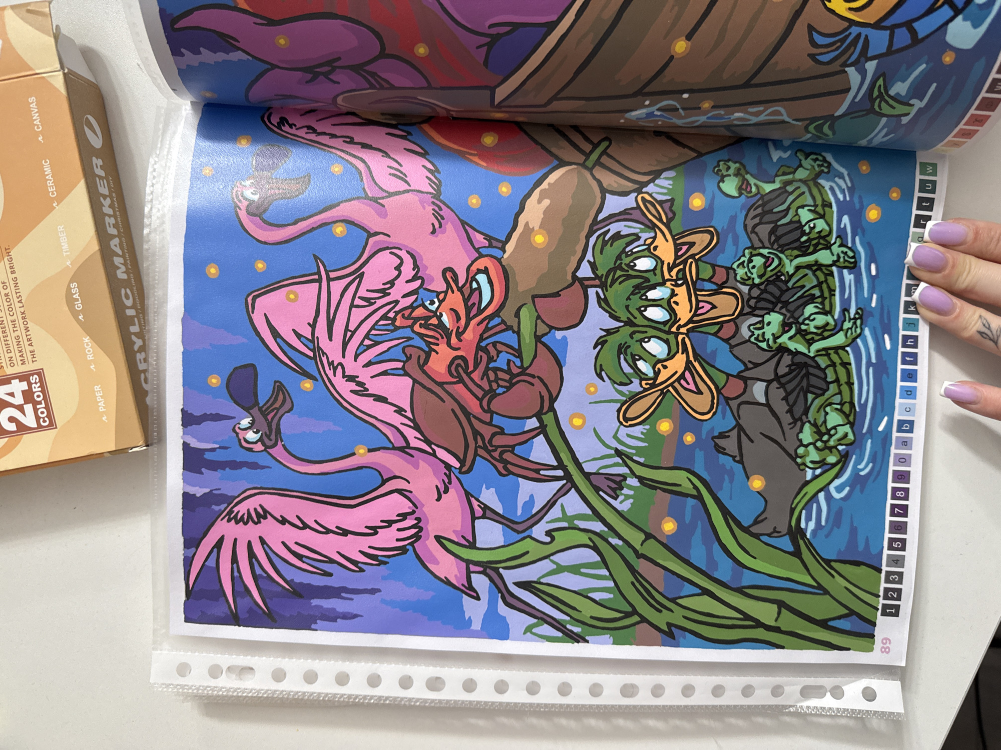 The image shows a page from a paint-by-numbers book featuring a cartoon illustration of flamingos, a crab, ducks, and turtles in a swampy environment. The illustration is colorful and detailed, with a blue sky, purple trees, and a dark blue body of water. There are also small yellow circles scattered throughout the image, possibly representing fireflies or stars. The bottom of the page has a numbered and lettered key for the paint-by-numbers activity. The book is open and placed on a white surface, with a box of "24 COLORS" acrylic markers visible in the upper right corner. A person's hand with painted fingernails is visible in the lower right corner, near the bottom of the page. The background suggests an indoor setting. There is no information in the image that indicates its location in Łódź, Poland.
